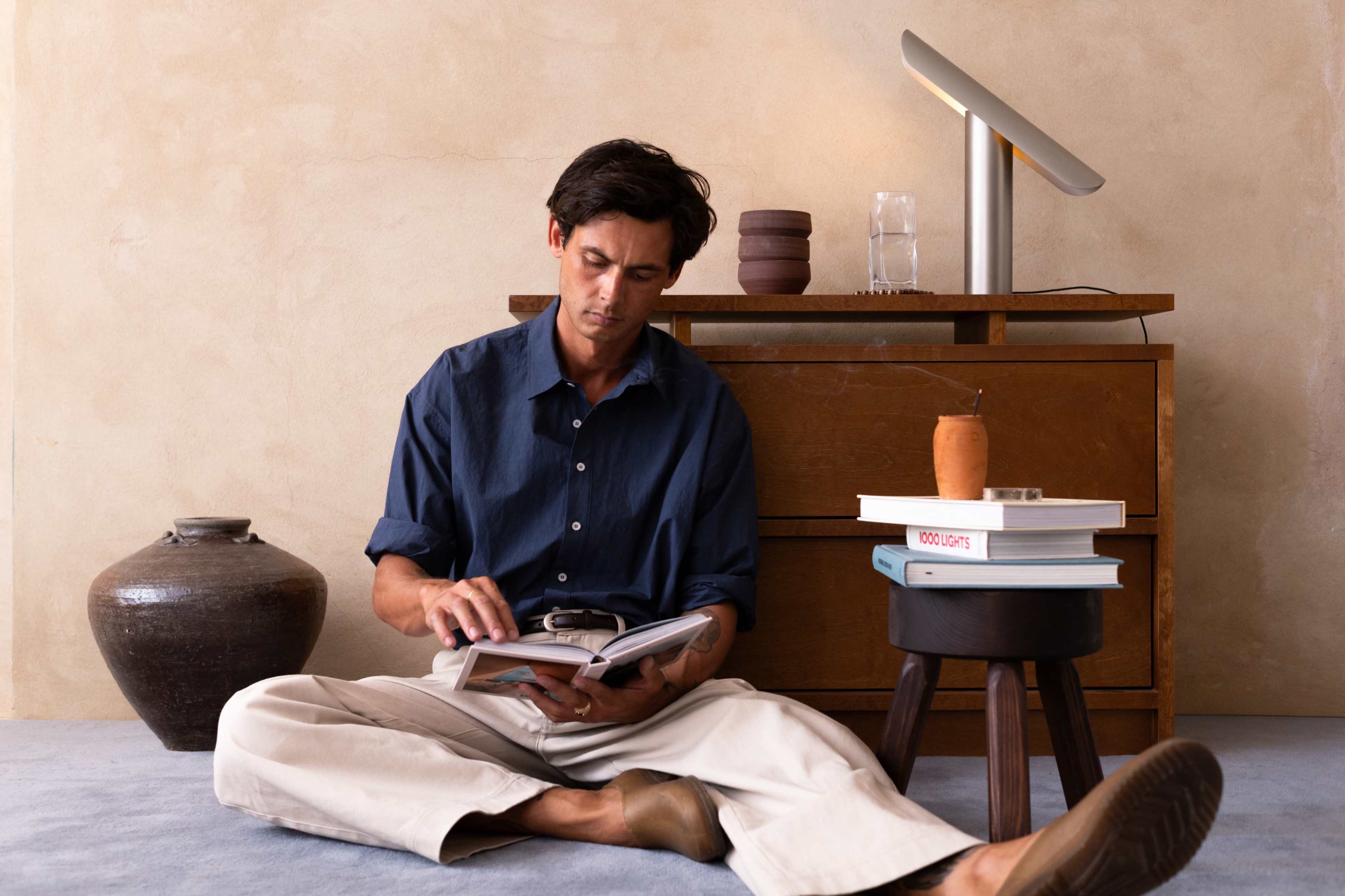 A man sits on the floor, reading a book beside a wooden cabinet that holds a stack of books and a ceramic pot.