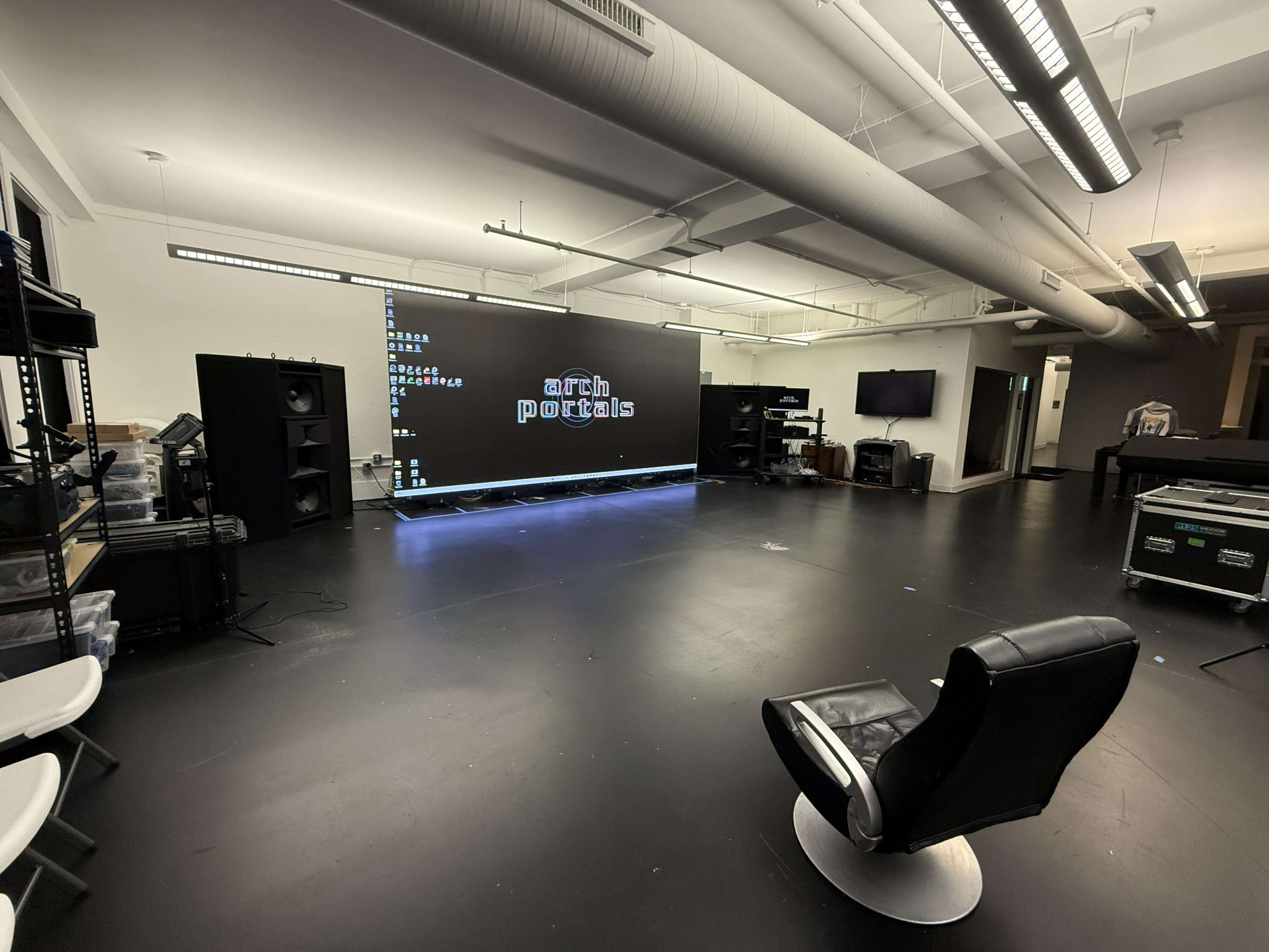 The image shows a spacious room equipped with audio equipment, a large screen displaying a text logo, and a single black chair in the center.