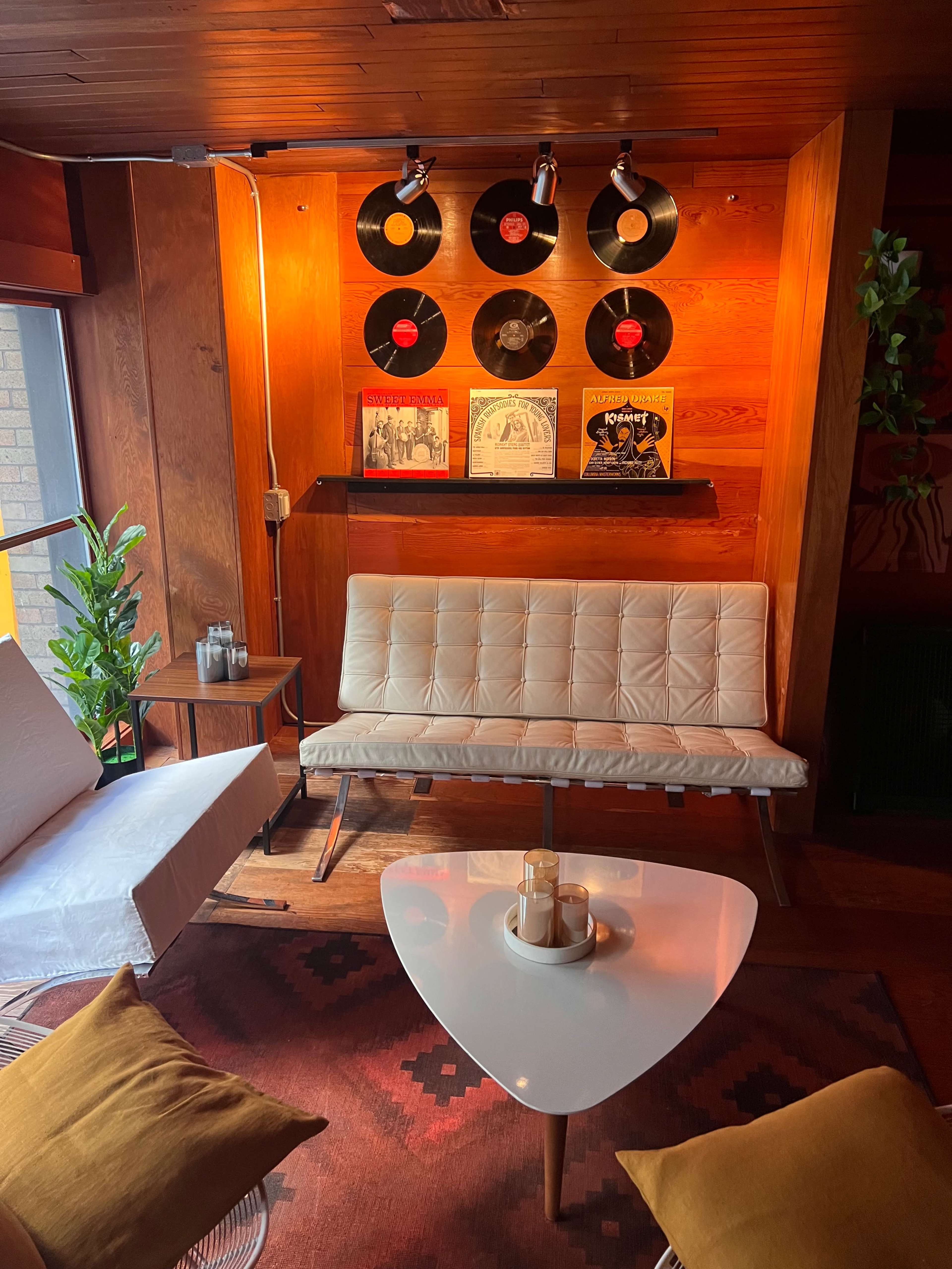 A cozy seating area with a white couch, a triangular coffee table, and vintage vinyl records displayed on a wooden wall.