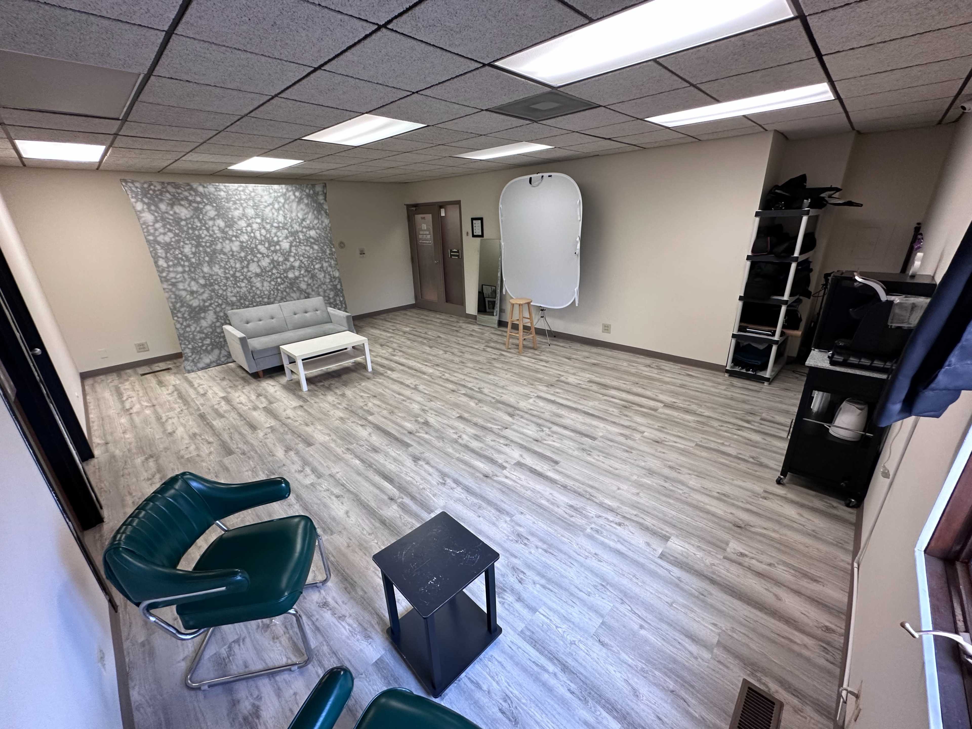 A spacious room with light gray walls, a hardwood floor, a gray sofa, a coffee table, and two green chairs, along with a photography backdrop and a light stand.