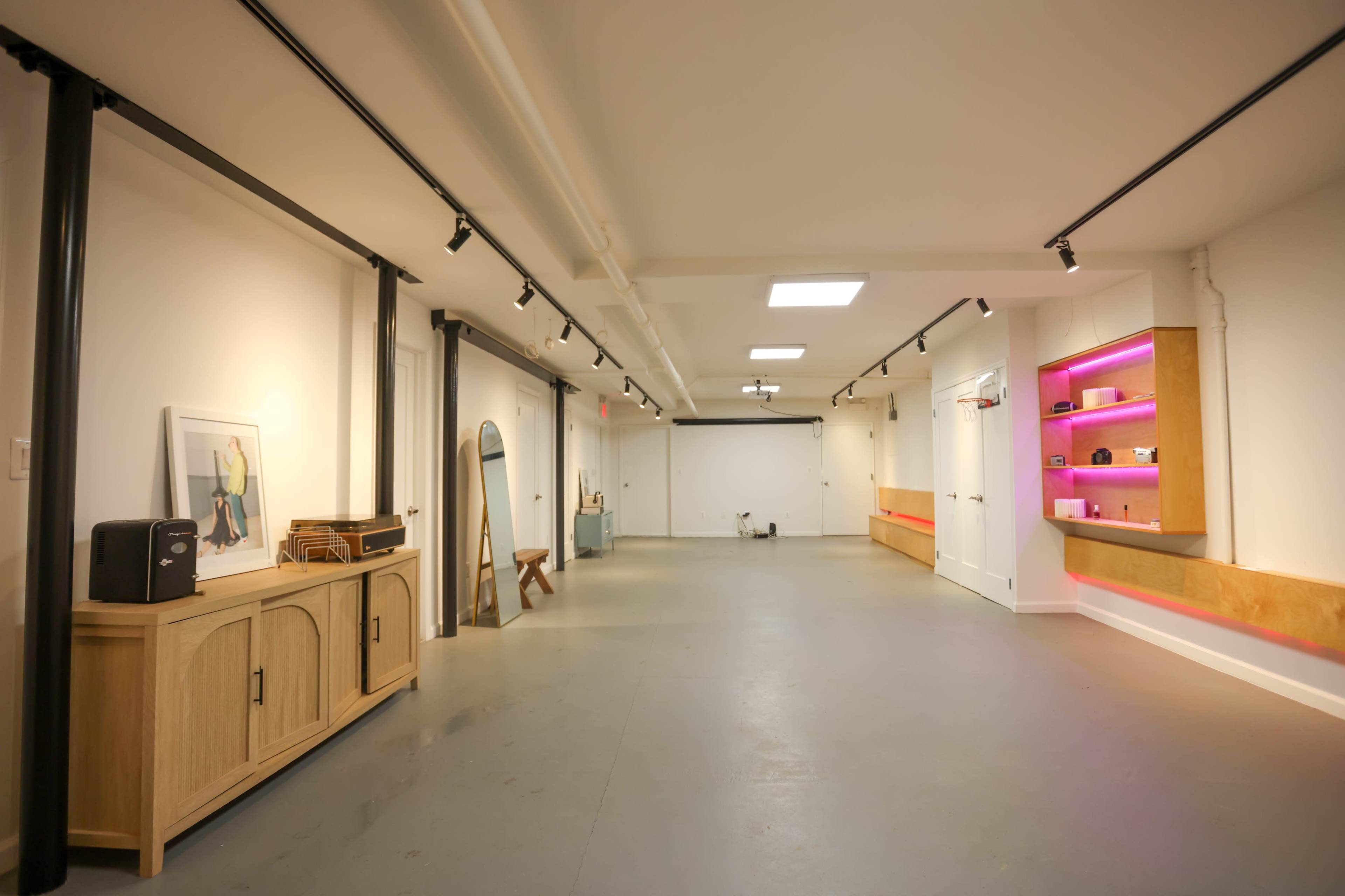 The image shows a spacious, well-lit interior featuring a polished concrete floor, white walls, and modern lighting, with a wooden cabinet and seating area along one side.