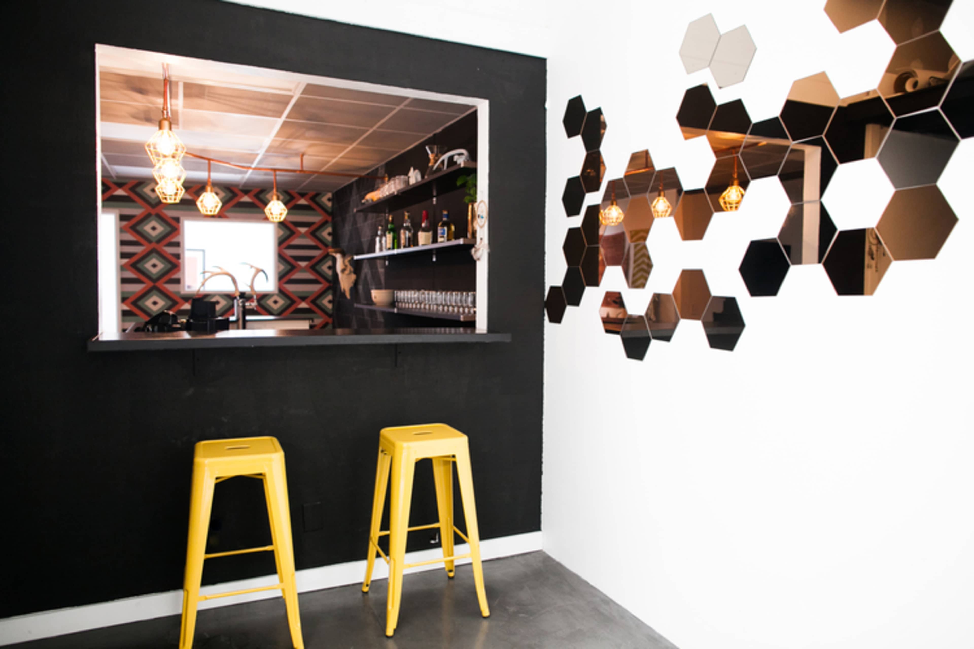 A bar area features a black wall with a window opening, two yellow stools in front, and hexagonal mirrors arranged decoratively on the adjacent white wall.