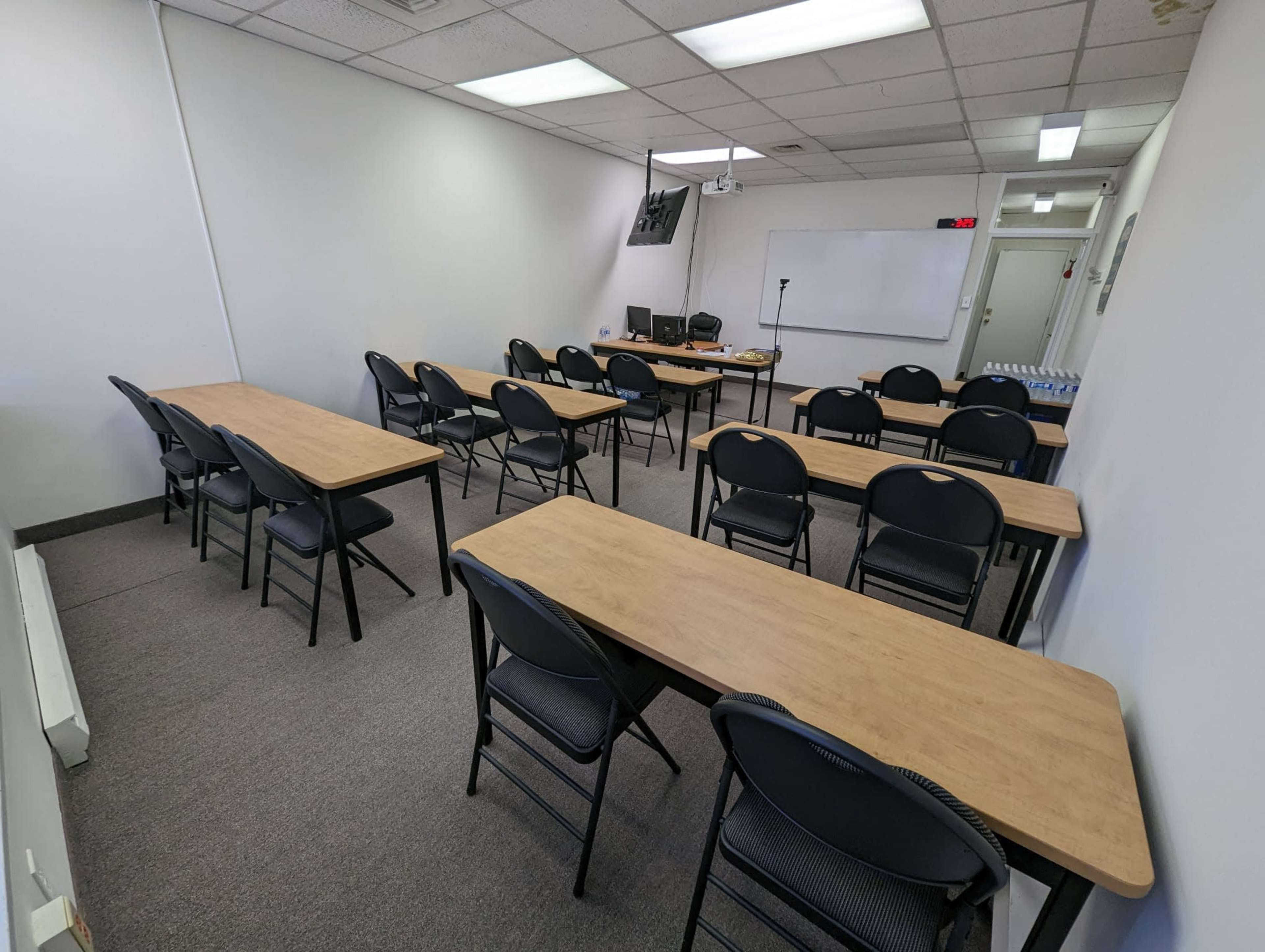 A classroom features several rows of tables and chairs arranged in an organized manner with a whiteboard and electronic equipment at the front.
