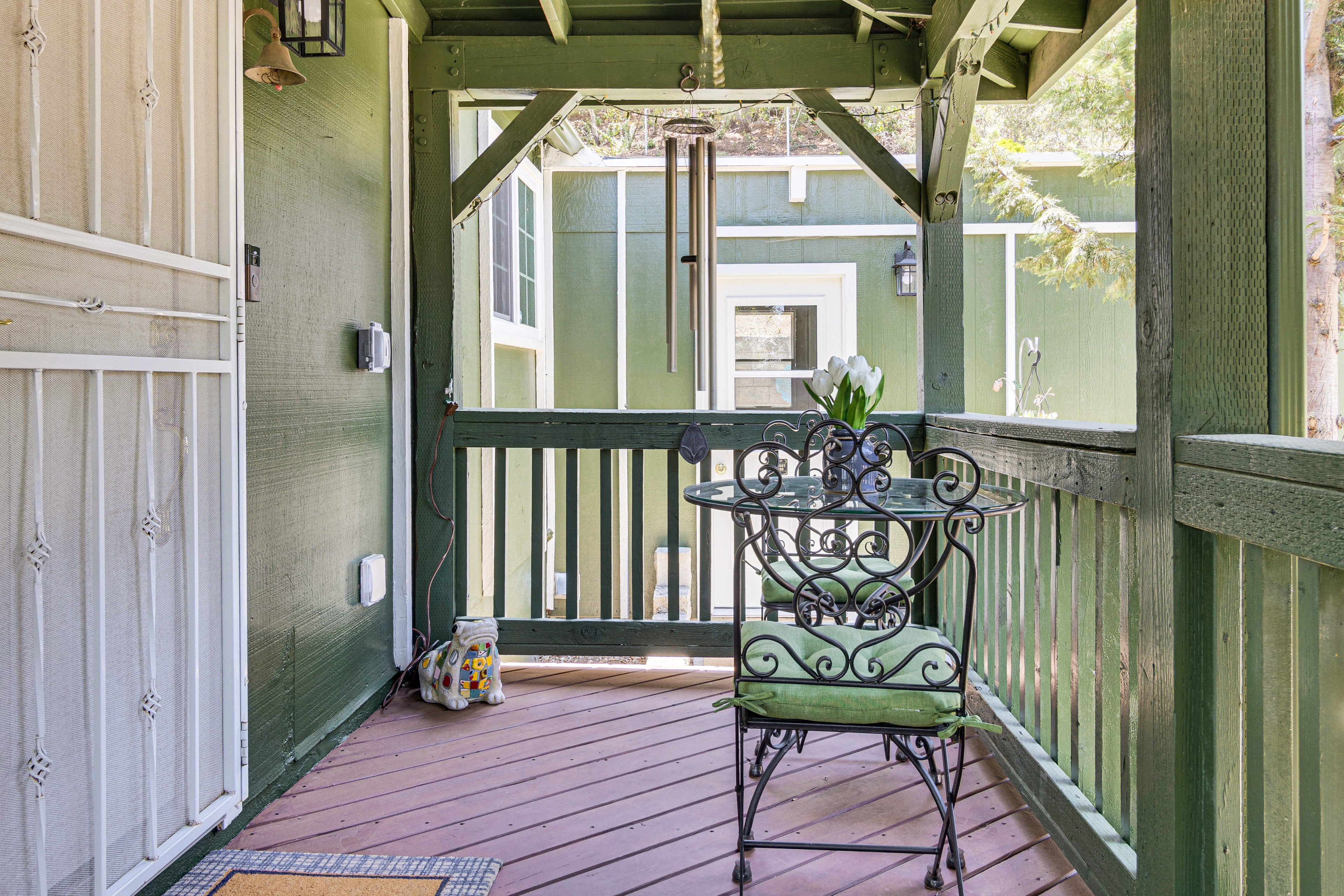 A green porch features a wrought iron table and chair set, adorned with a vase of tulips, and is framed by wooden railings and wind chimes.