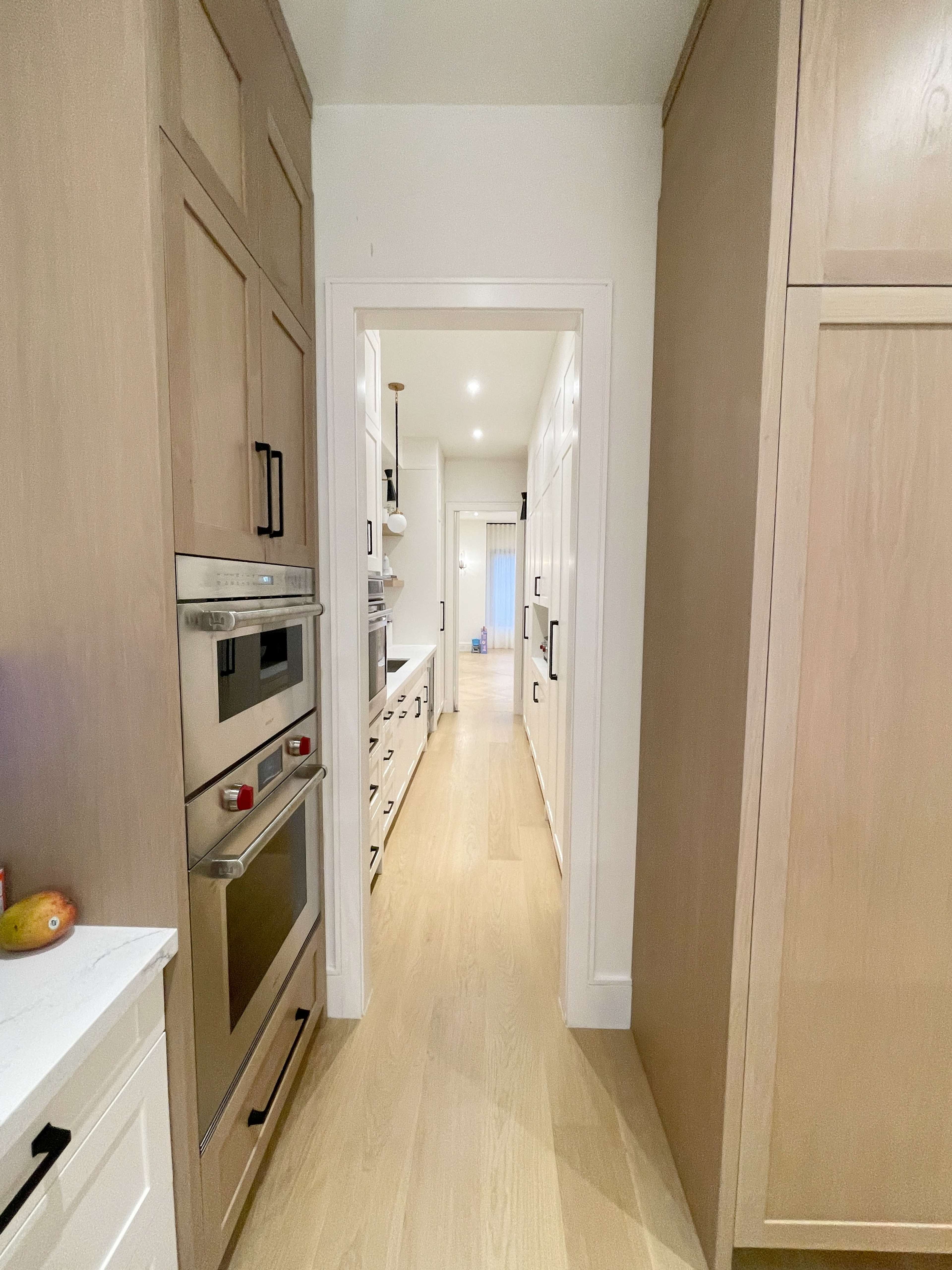 A narrow hallway connects two sections of a modern kitchen, featuring cabinetry on both sides and appliances along the left wall.