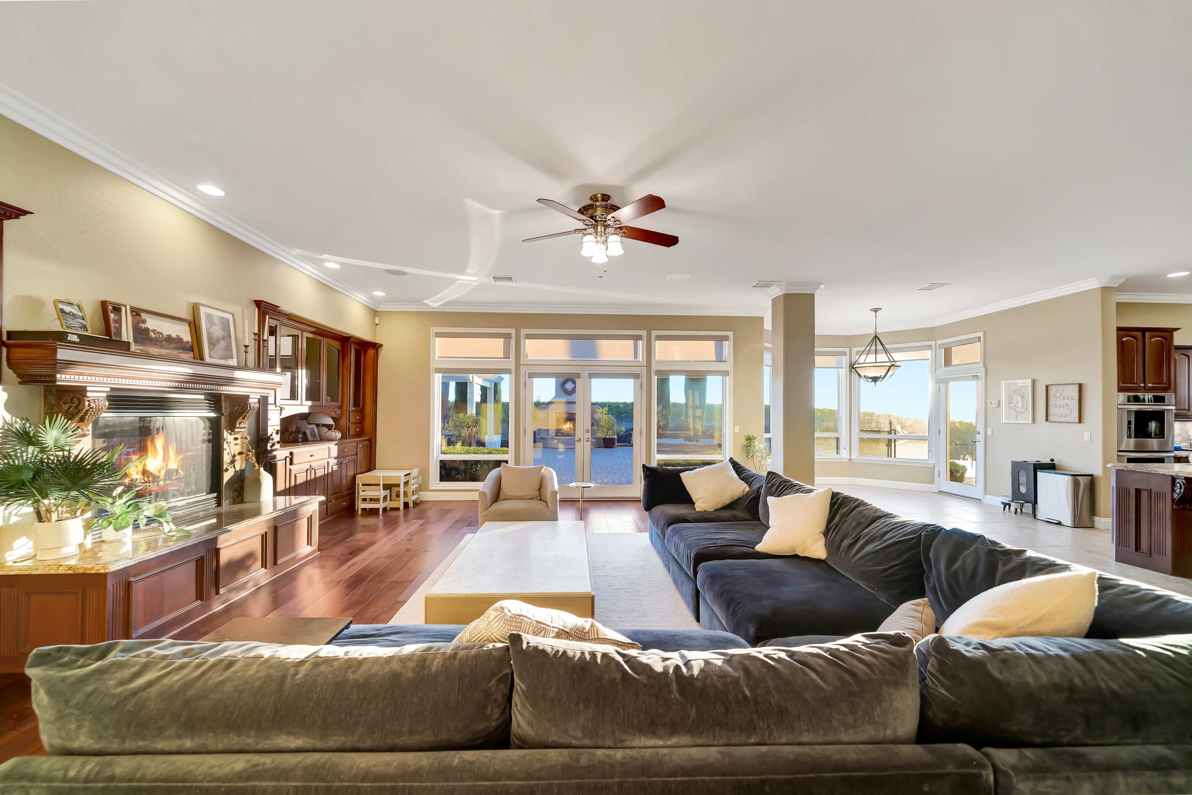 The image shows a spacious, well-lit living room featuring a large sectional sofa, a fireplace, and large windows overlooking a scenic view.