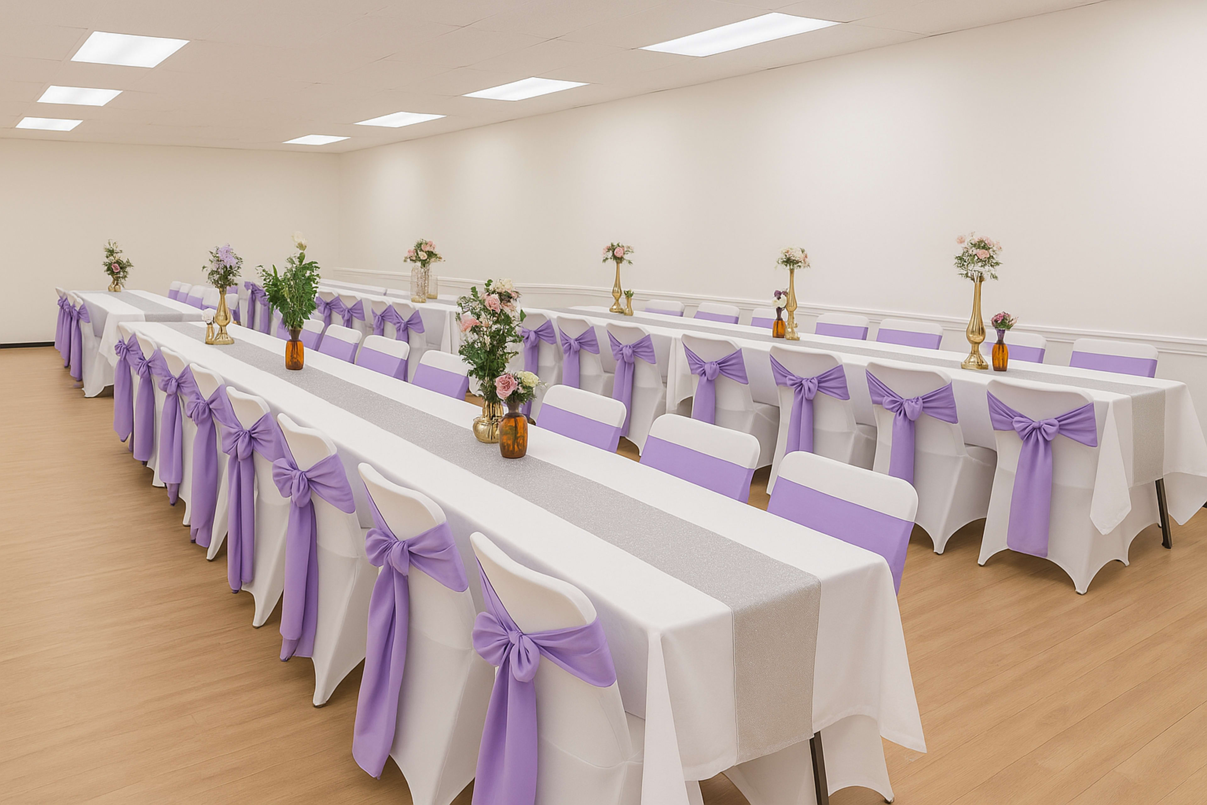 The image depicts a large, well-lit banquet hall set with multiple tables covered in white tablecloths, featuring lavender sashes and small floral centerpieces.