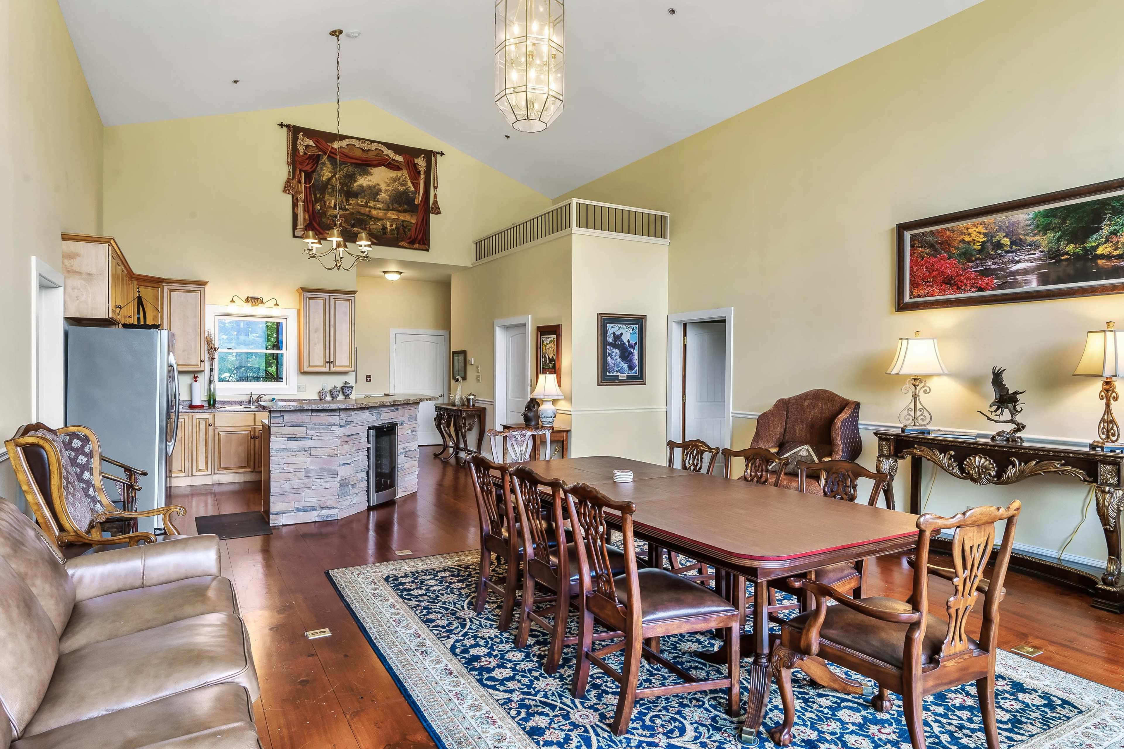 A spacious living area with a high ceiling, featuring a dining table surrounded by chairs, a sofa, and a kitchen area in the background.