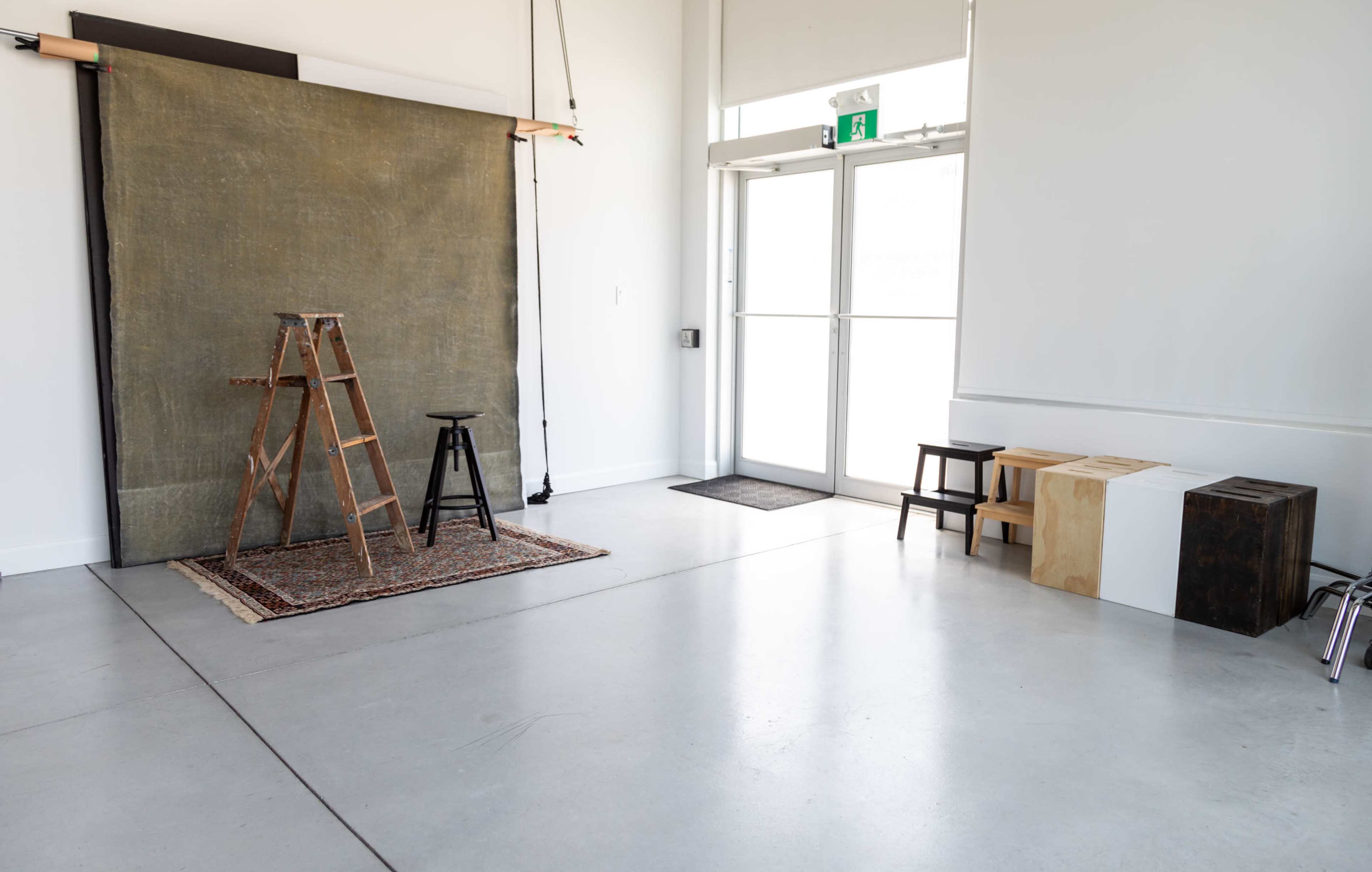 A photography studio featuring a wooden ladder, a stool, a backdrop draped on a frame, and various seating blocks arranged against a wall.