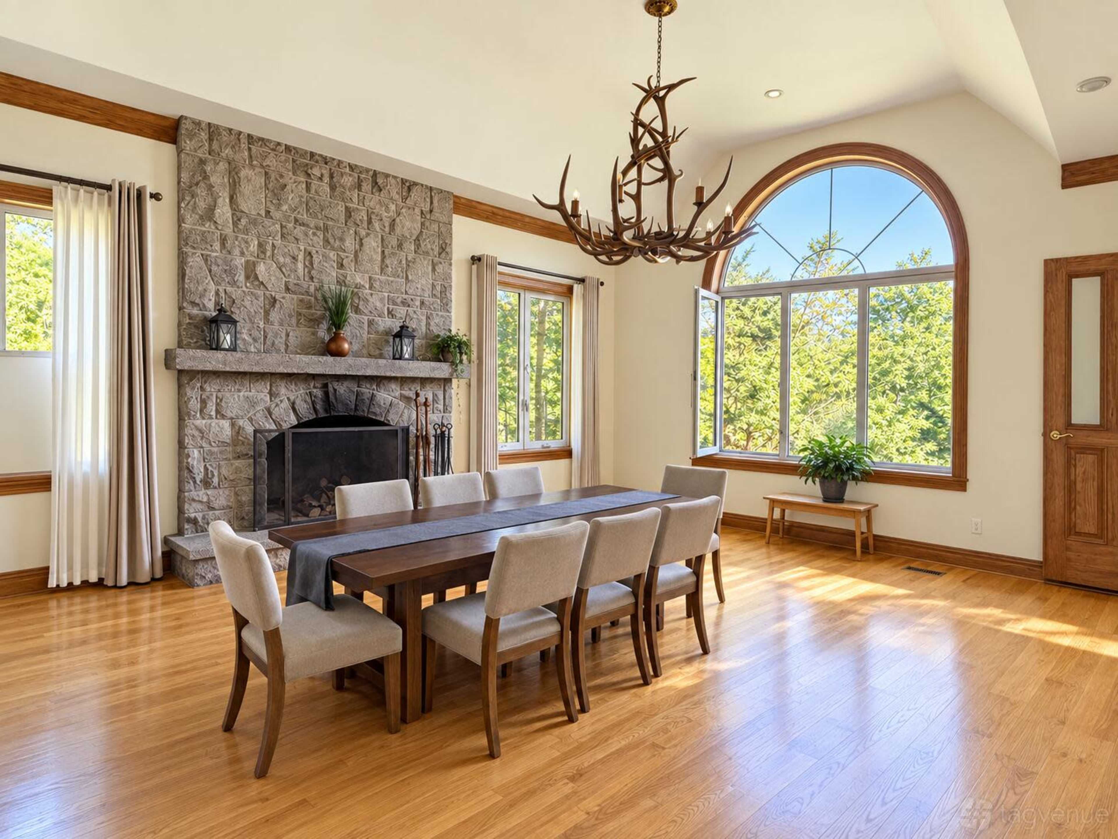 The image shows a spacious dining room with a long wooden table surrounded by beige chairs, a stone fireplace, and large windows letting in natural light.