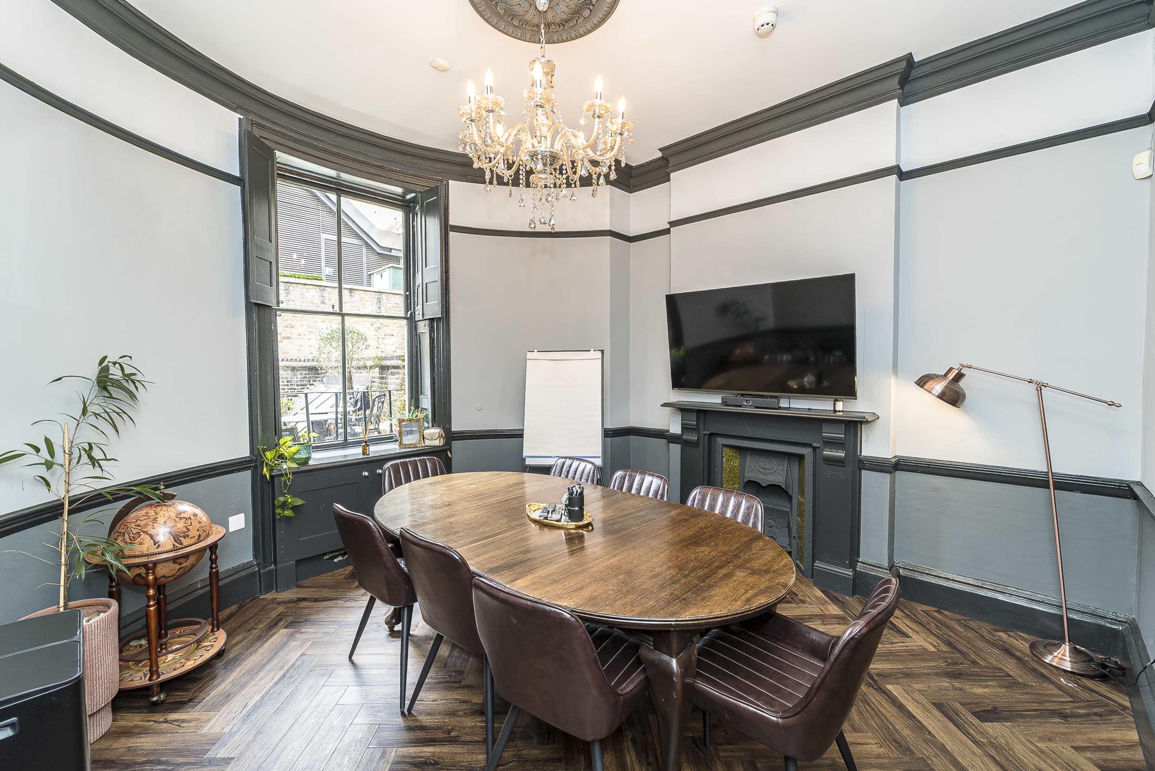 The image shows a well-appointed meeting room featuring a round wooden table surrounded by brown leather chairs, a large wall-mounted TV, and a chandelier hanging from the ceiling, with windows allowing natural light.
