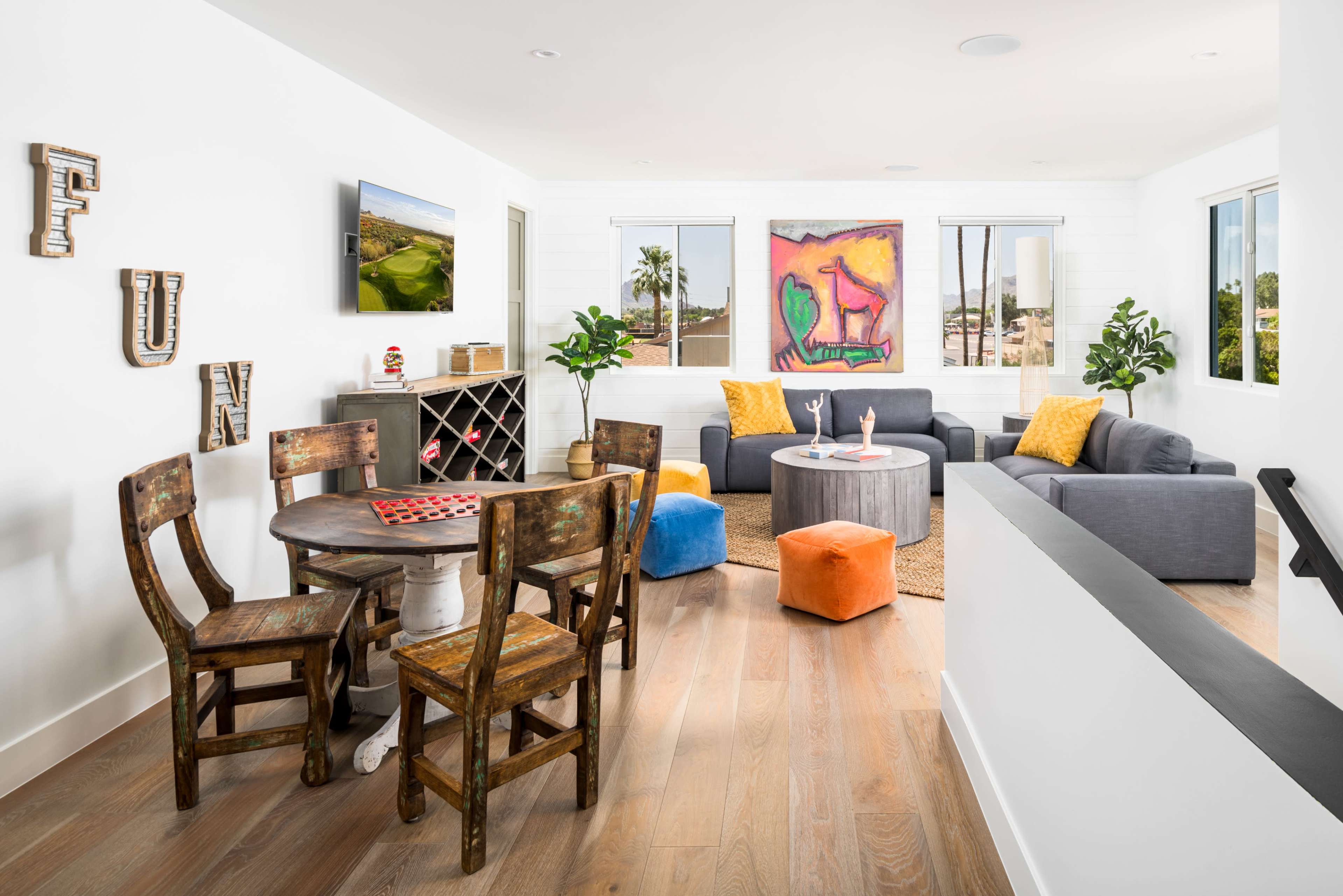 A bright, modern living area features a circular dining table with rustic chairs, a gray sofa set, colorful ottomans, and a large abstract painting on the wall.