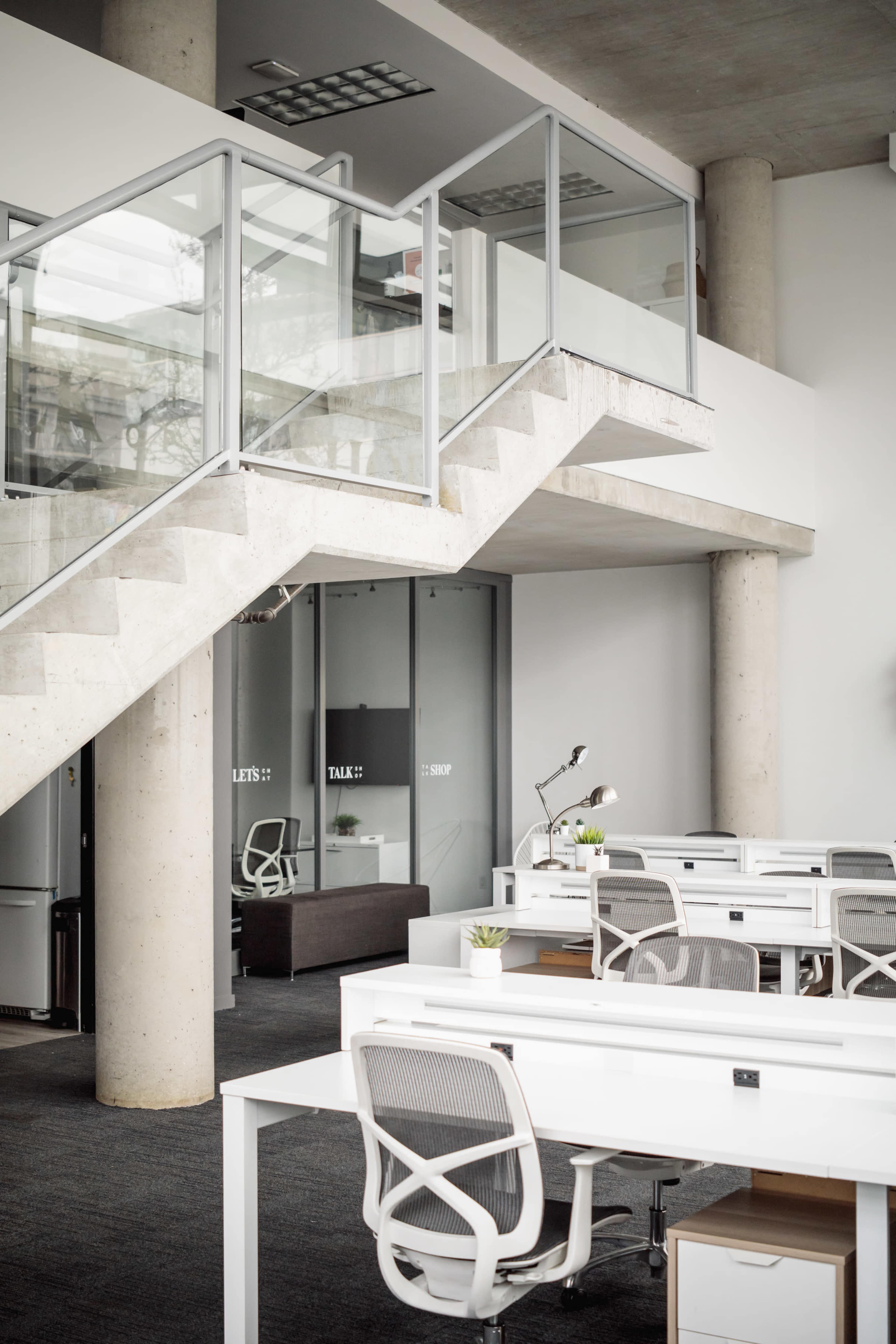 A modern office space featuring a staircase, glass partitions, and several white desks arranged throughout the area.