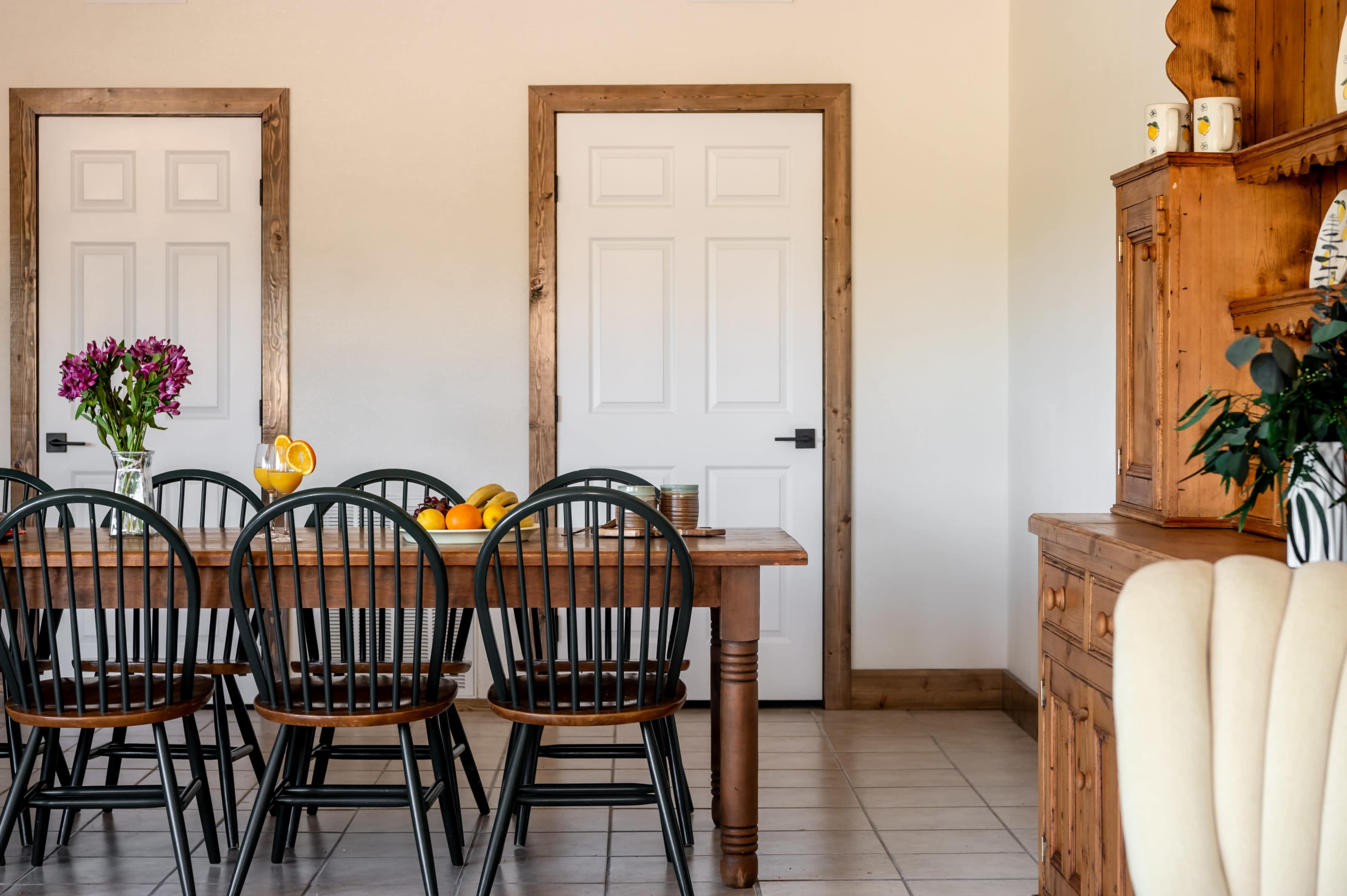 A wooden table with black chairs is set against a wall with two mirrors and a closed white door, alongside a wooden cabinet with decorative items.