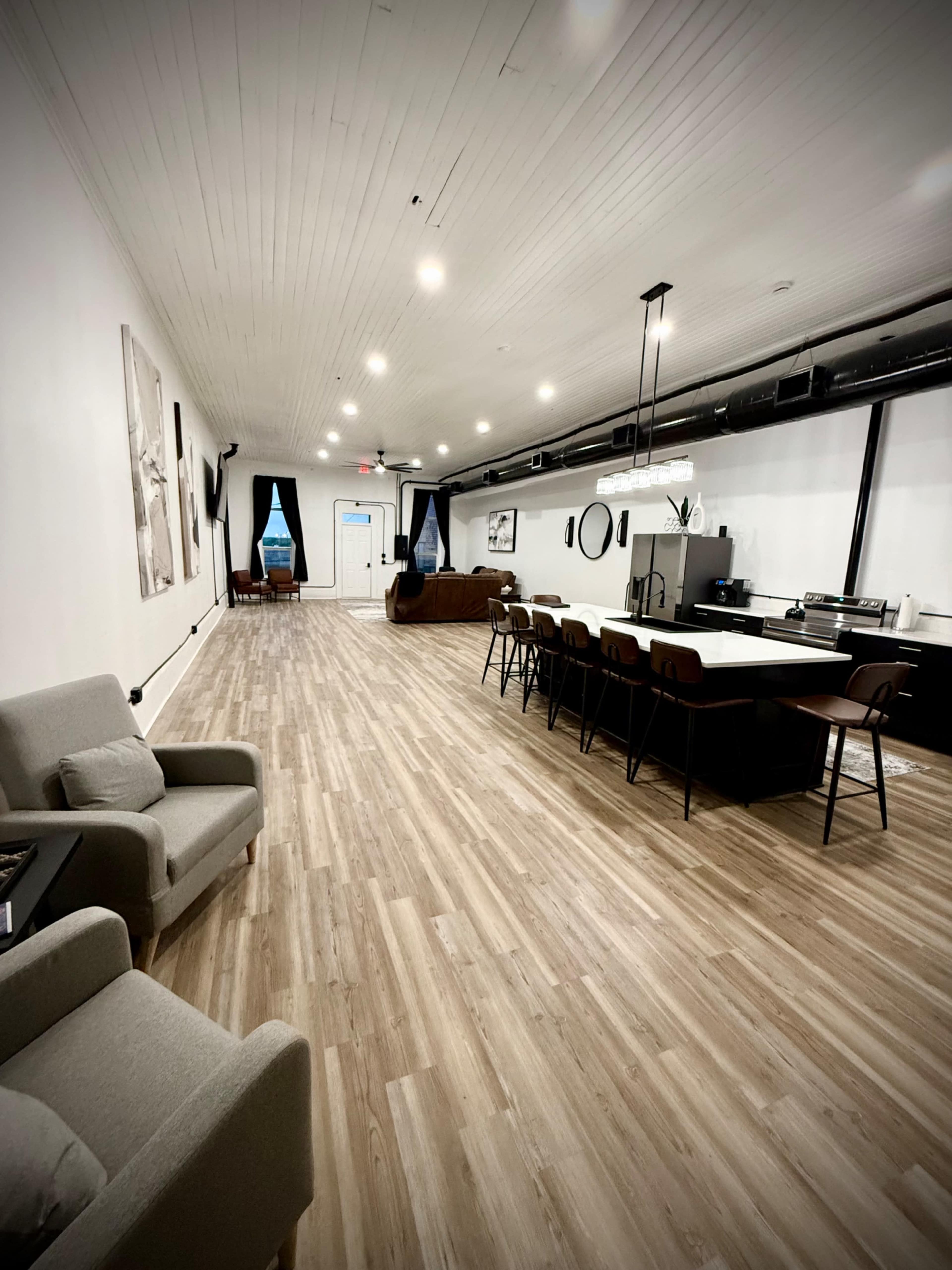 The image shows a modern, spacious room with wooden flooring, featuring a kitchen area on one side and a seating area with a gray armchair on the other.