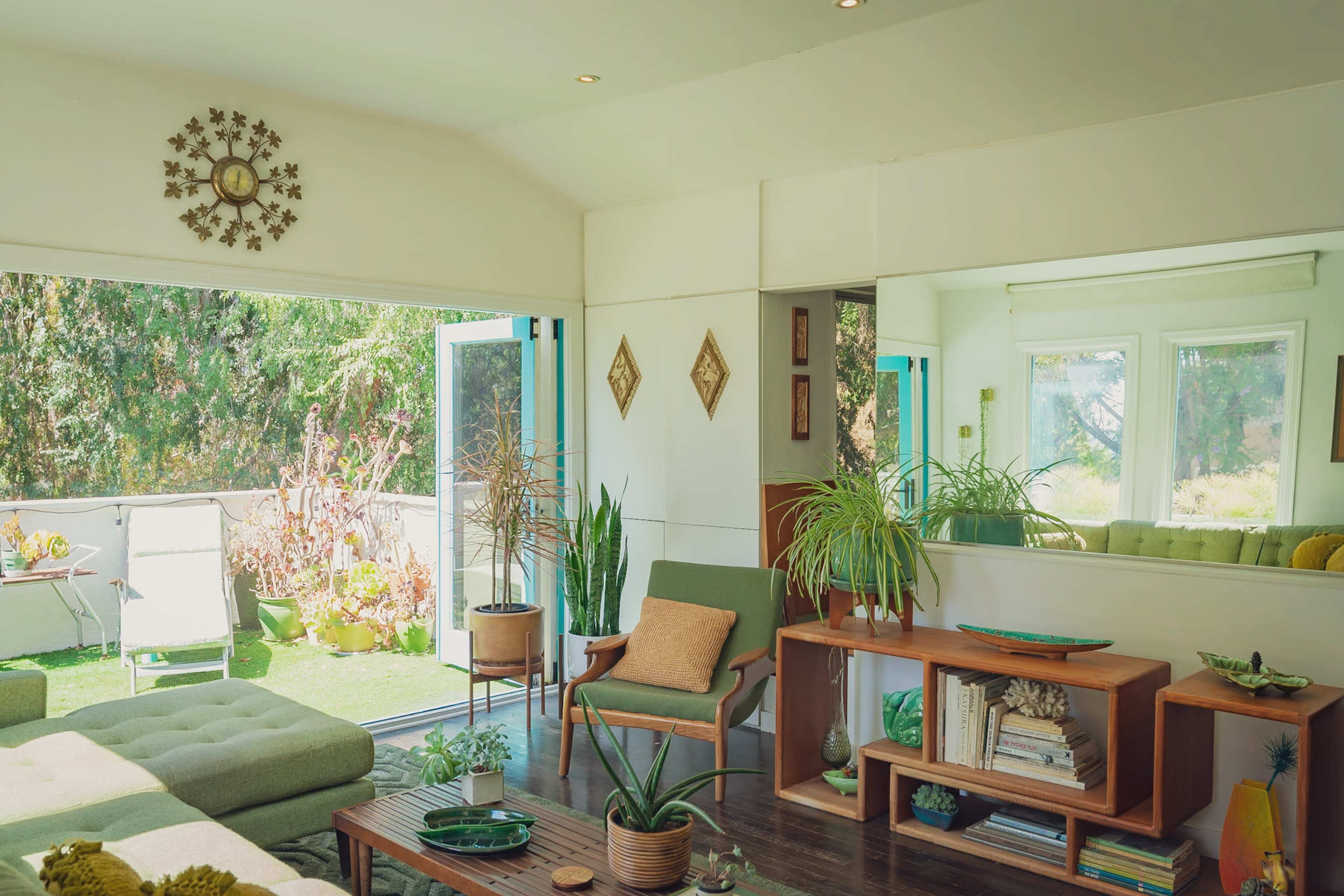 A bright living room features a green couch, wooden furniture, and large windows that open to a patio with plants.