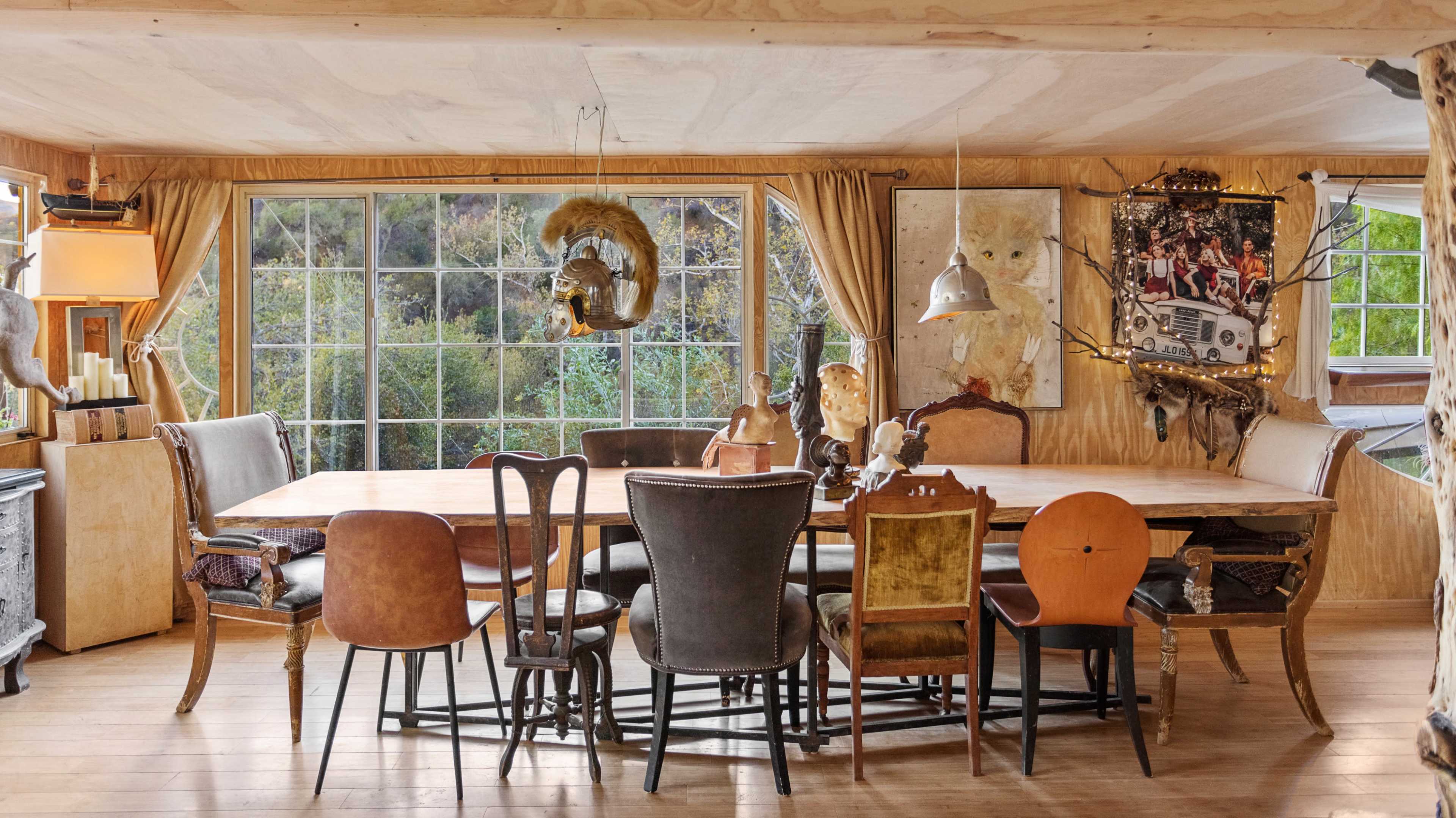 A dining room features a large wooden table surrounded by various chairs and windows that overlook a natural landscape.