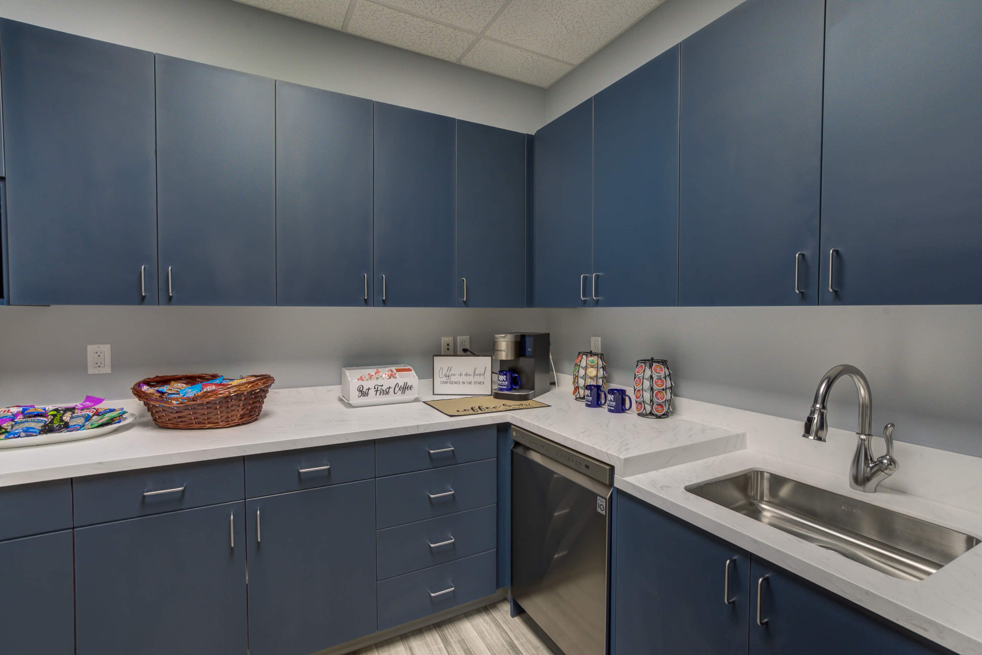 A modern kitchen with blue cabinets, a stainless steel dishwasher, a sink, and a countertop displaying a basket and decorative mugs.