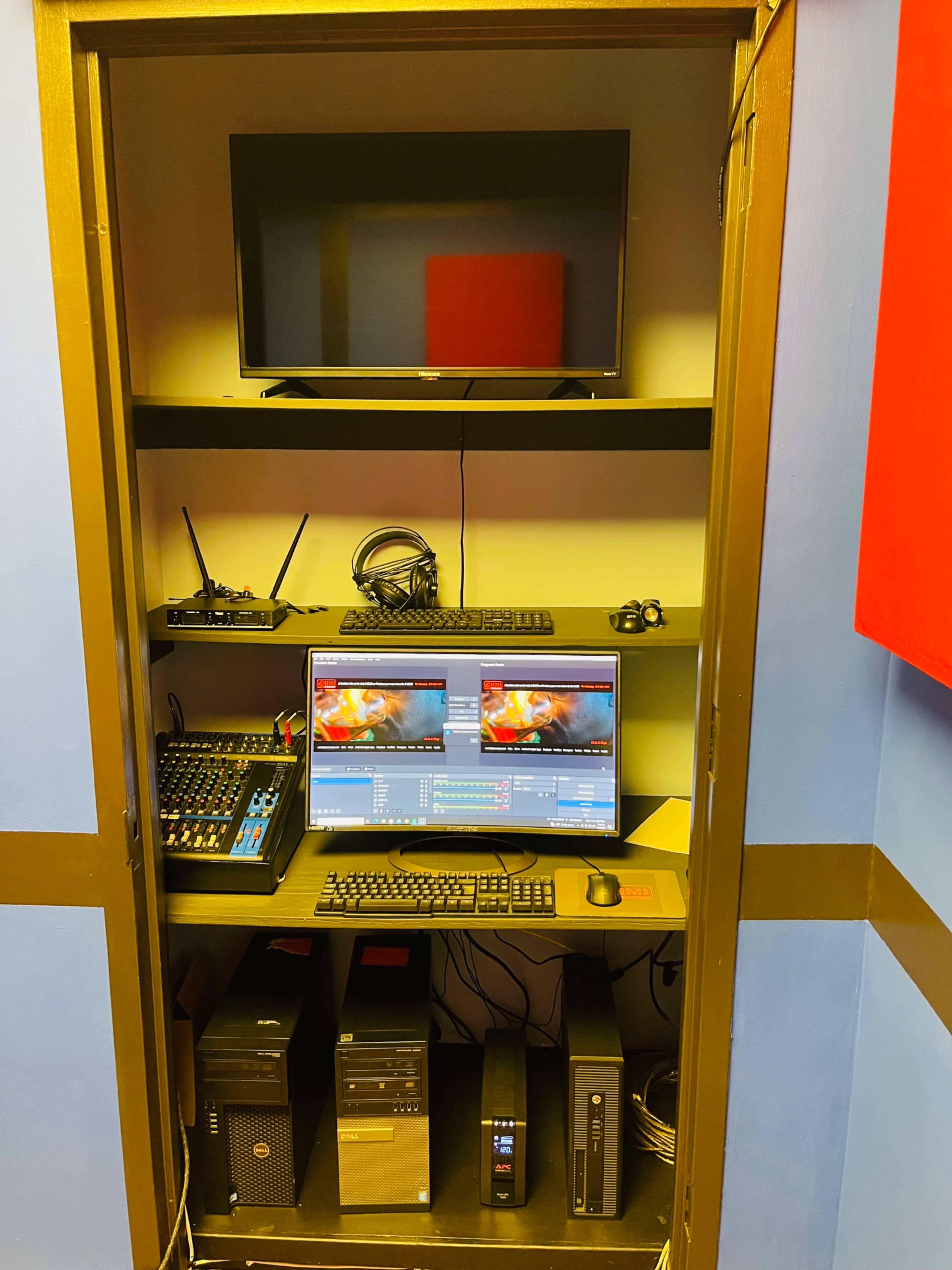 A compact workspace features two computer monitors, an audio mixer, a television, and various tech equipment organized on shelves.
