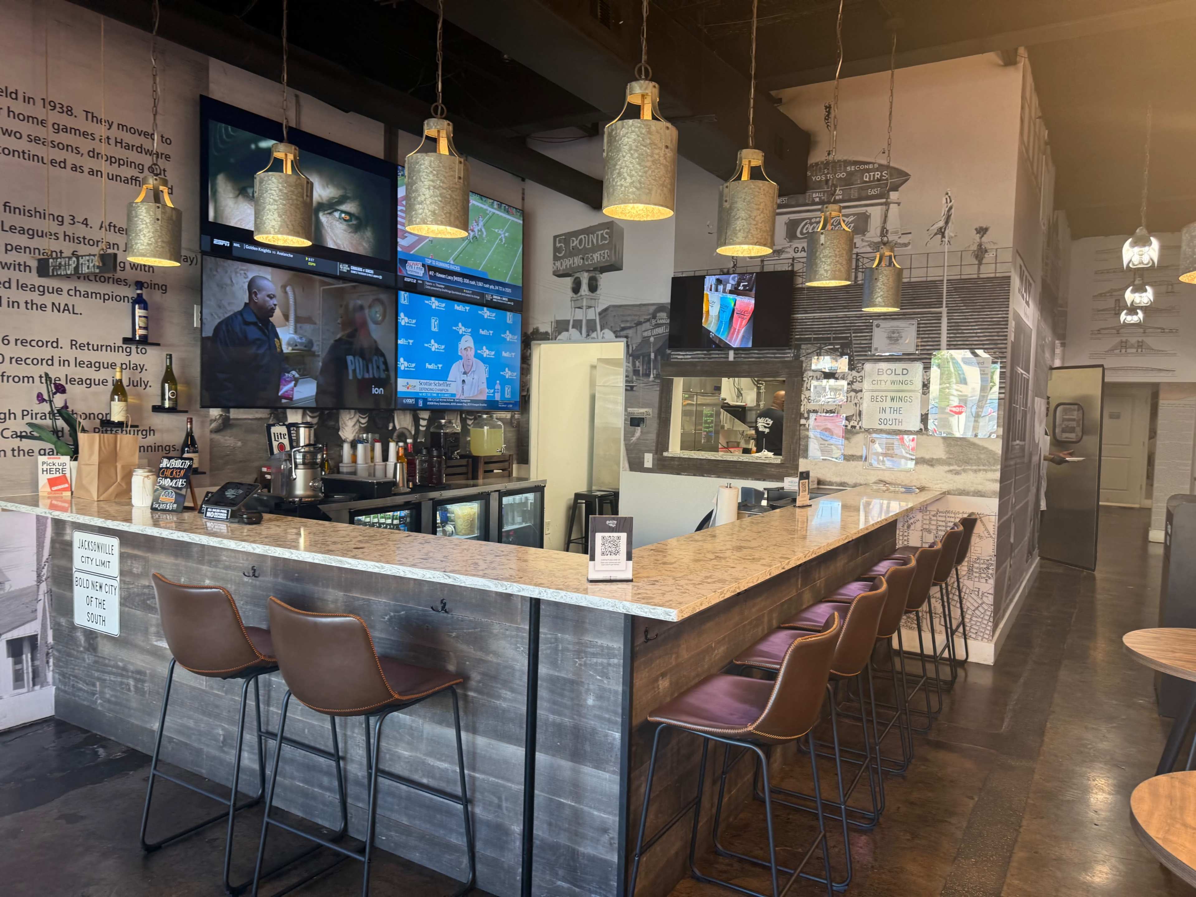 The image shows a modern bar area with a granite countertop, several high stools, and multiple screens displaying sports content.