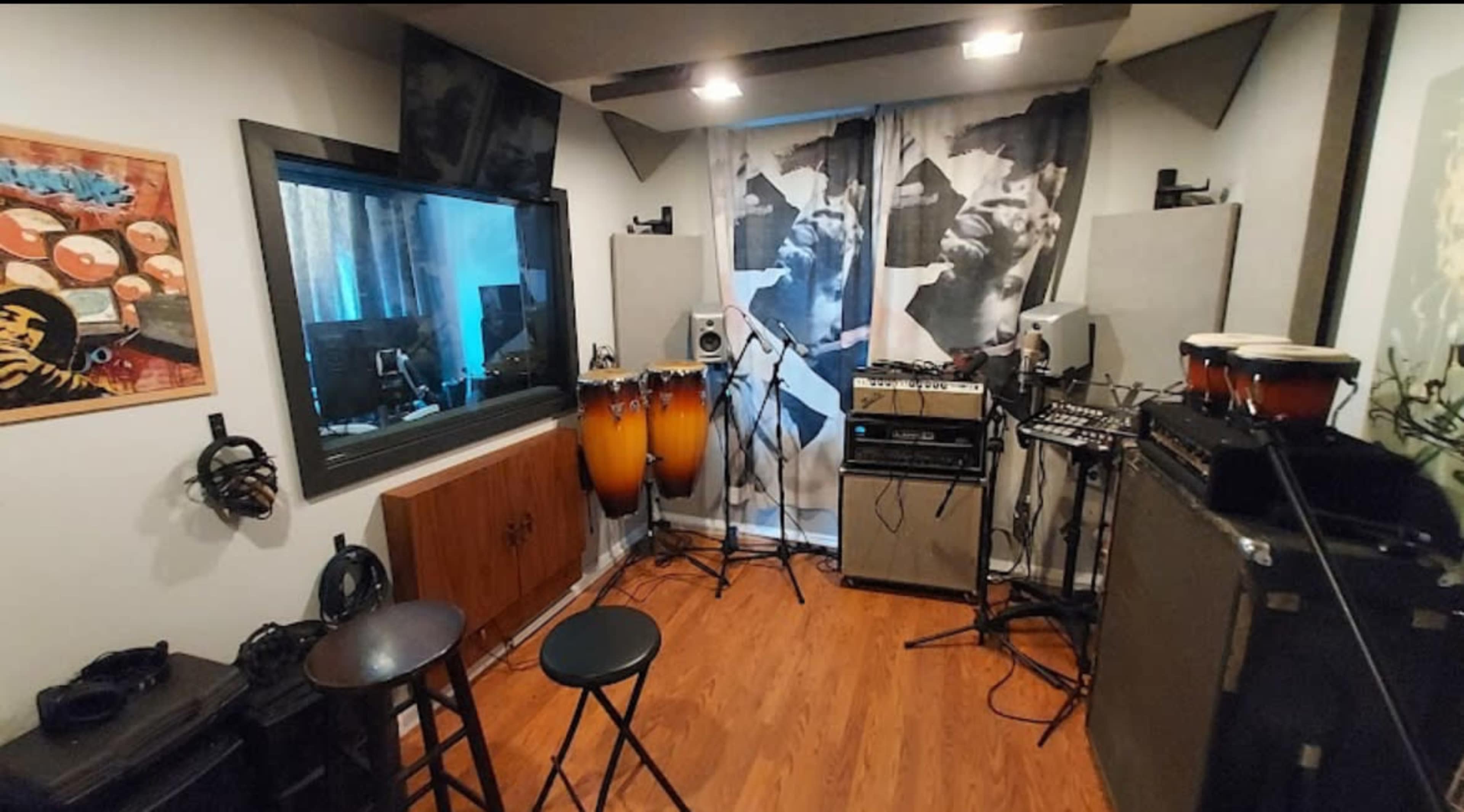 The image shows a music studio with various instruments, including congas, an amplifier, and recording equipment, set against a backdrop of patterned curtains.