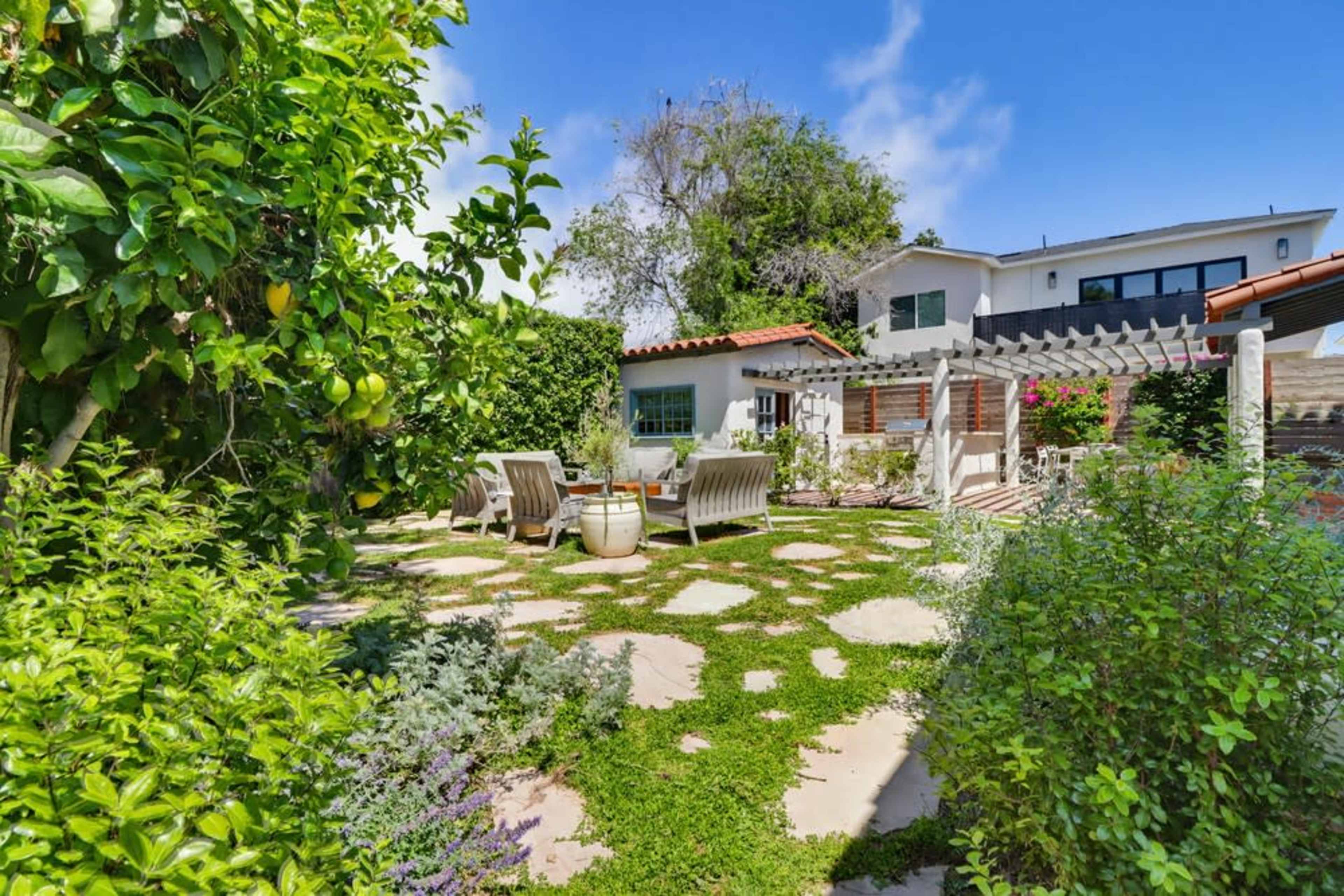 The image shows a landscaped outdoor garden area featuring a stone path, a seating arrangement, and greenery surrounding a house.
