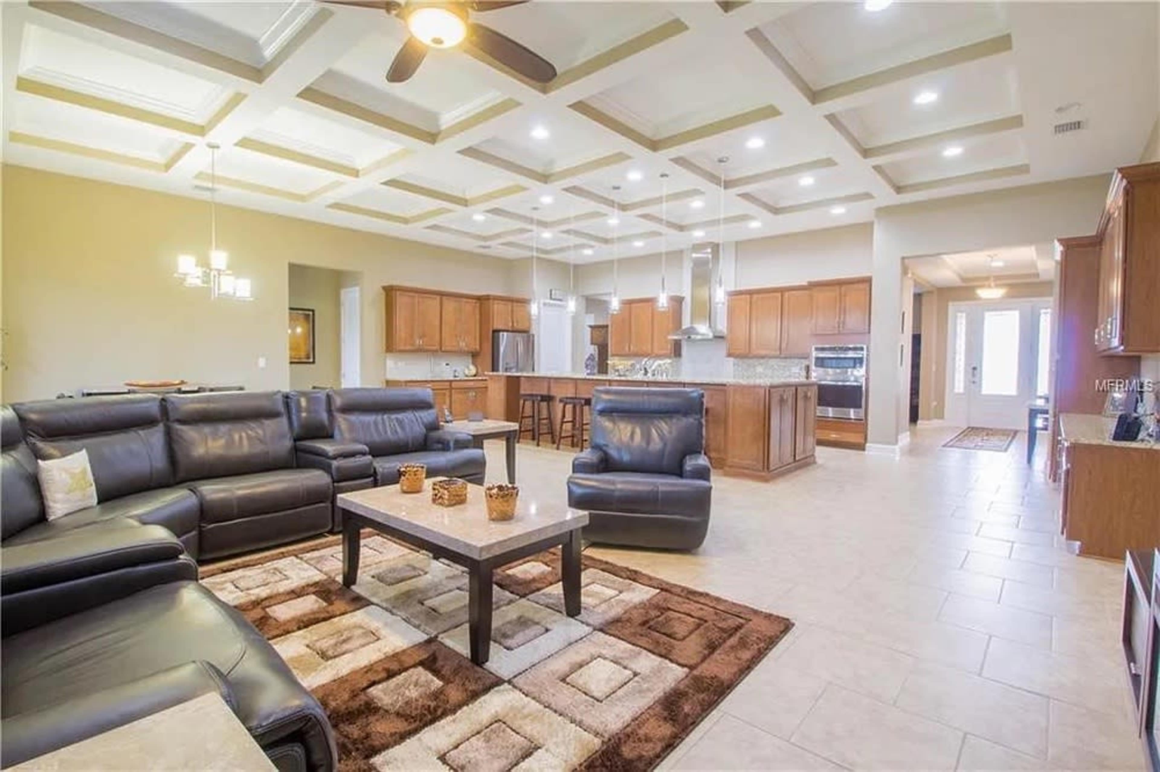 The image shows a spacious, modern living room with a coffered ceiling, dark leather furniture, and an open layout connecting to a kitchen area.