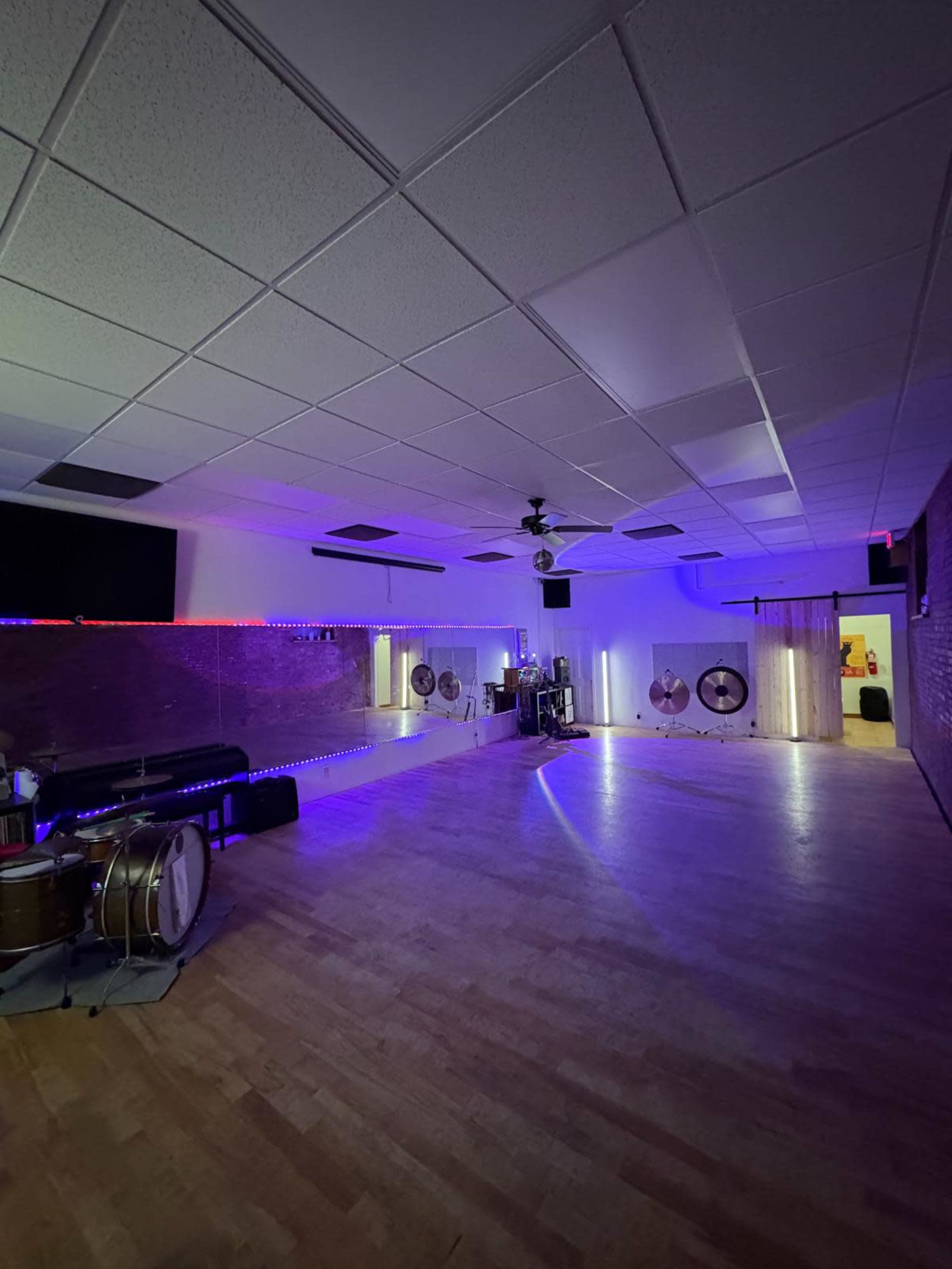 The image shows a spacious, well-lit room with wooden flooring, featuring various musical instruments, neon lights, and reflective surfaces on the walls.