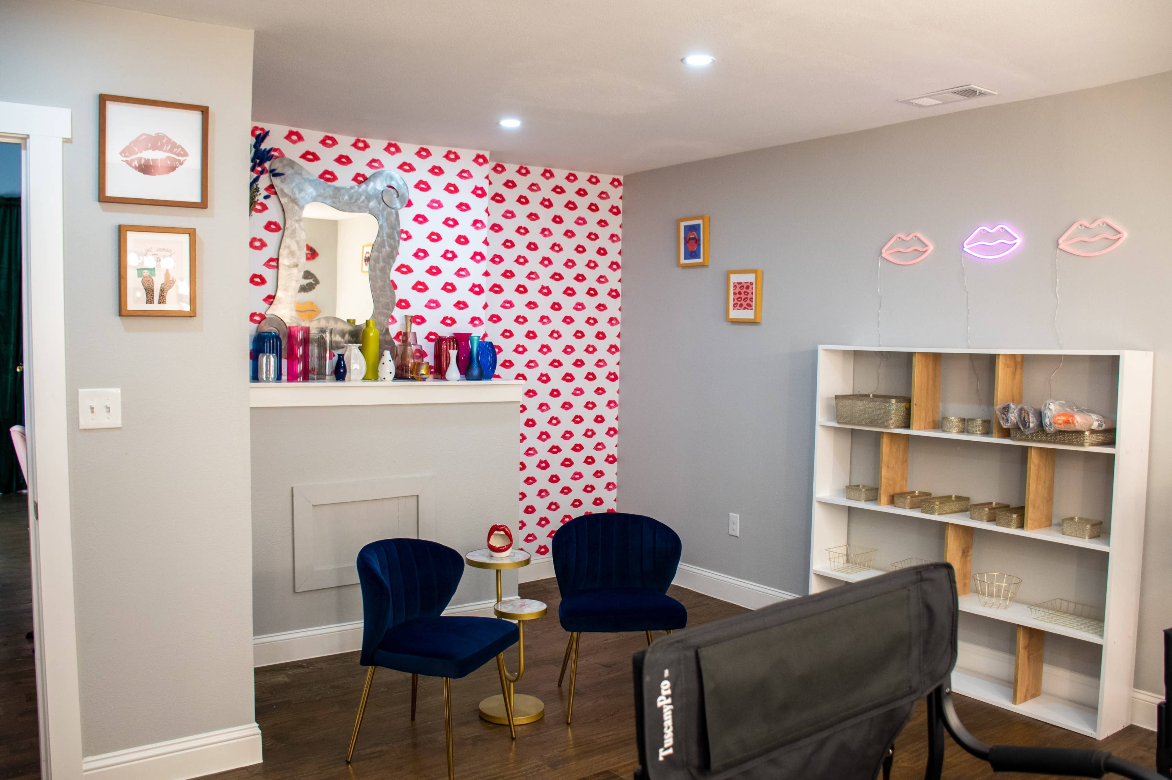 A cozy room with two blue chairs and a small white shelf, decorated with a red lip-patterned wall and various colorful items on a mantle.