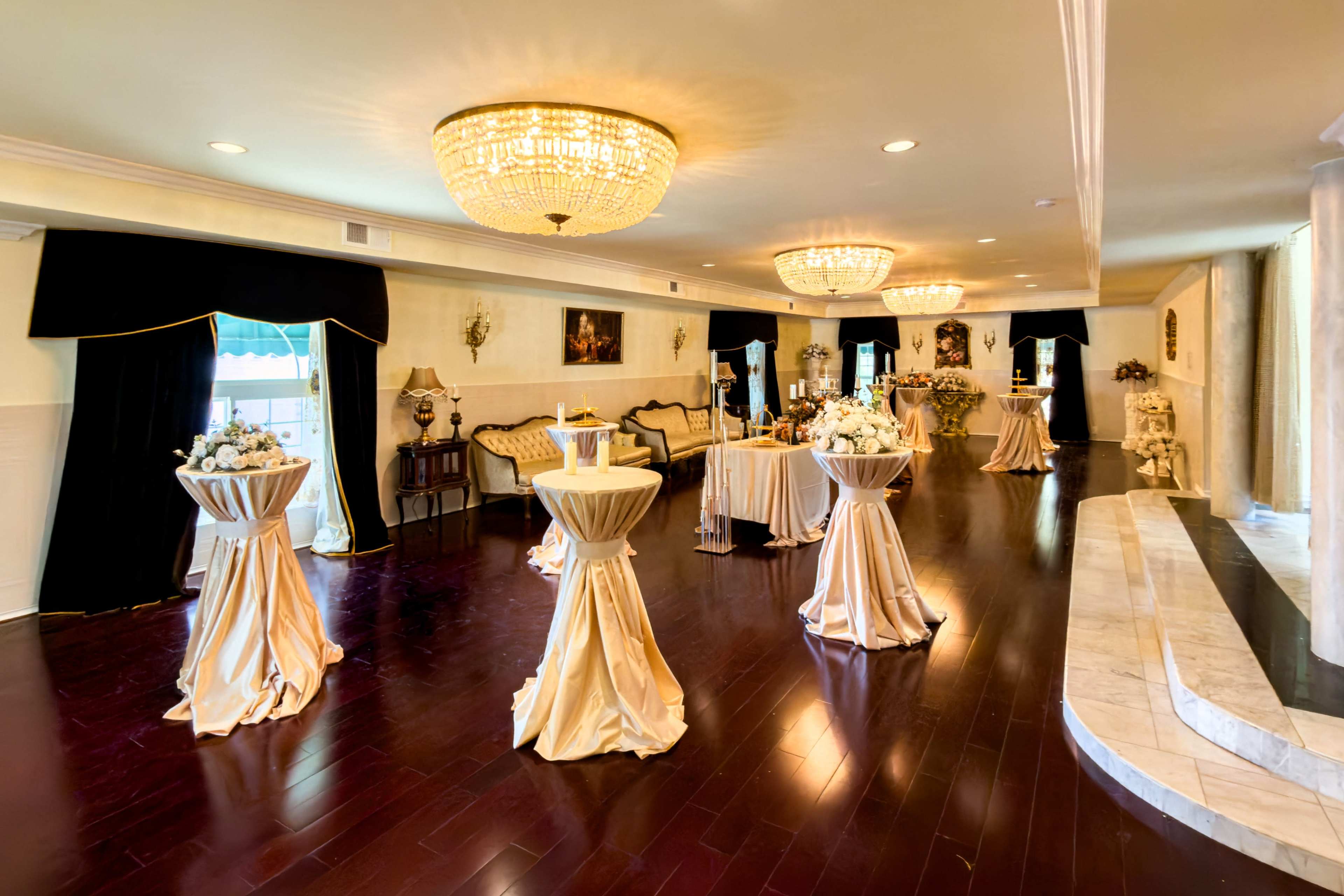 The image shows a spacious event room with polished dark wooden floors, elegantly draped tables, and chandeliers providing overhead lighting.