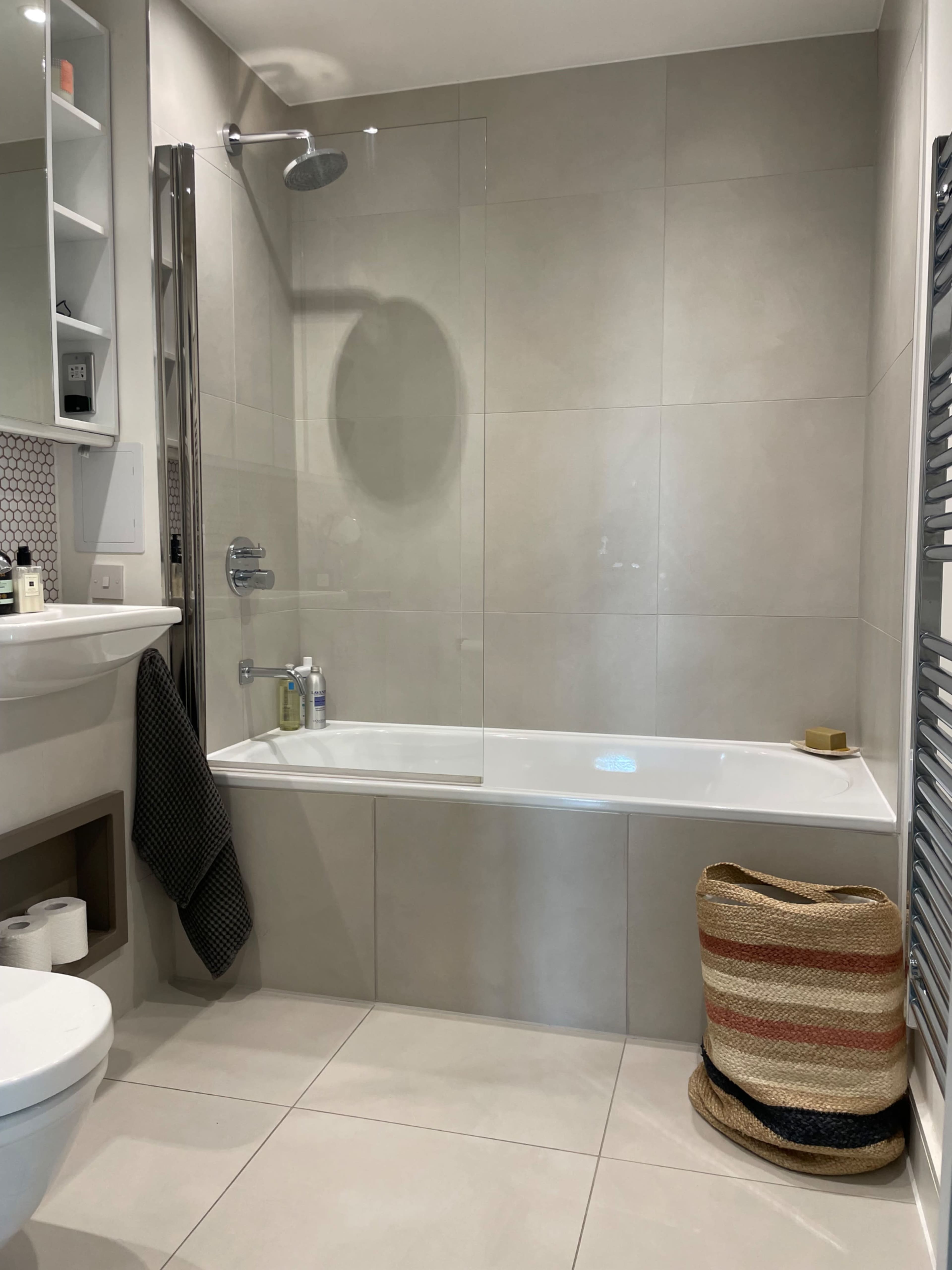 A modern bathroom features a bathtub with a glass shower screen, a wall-mounted mirror with shelves, and a woven basket beside the tub.