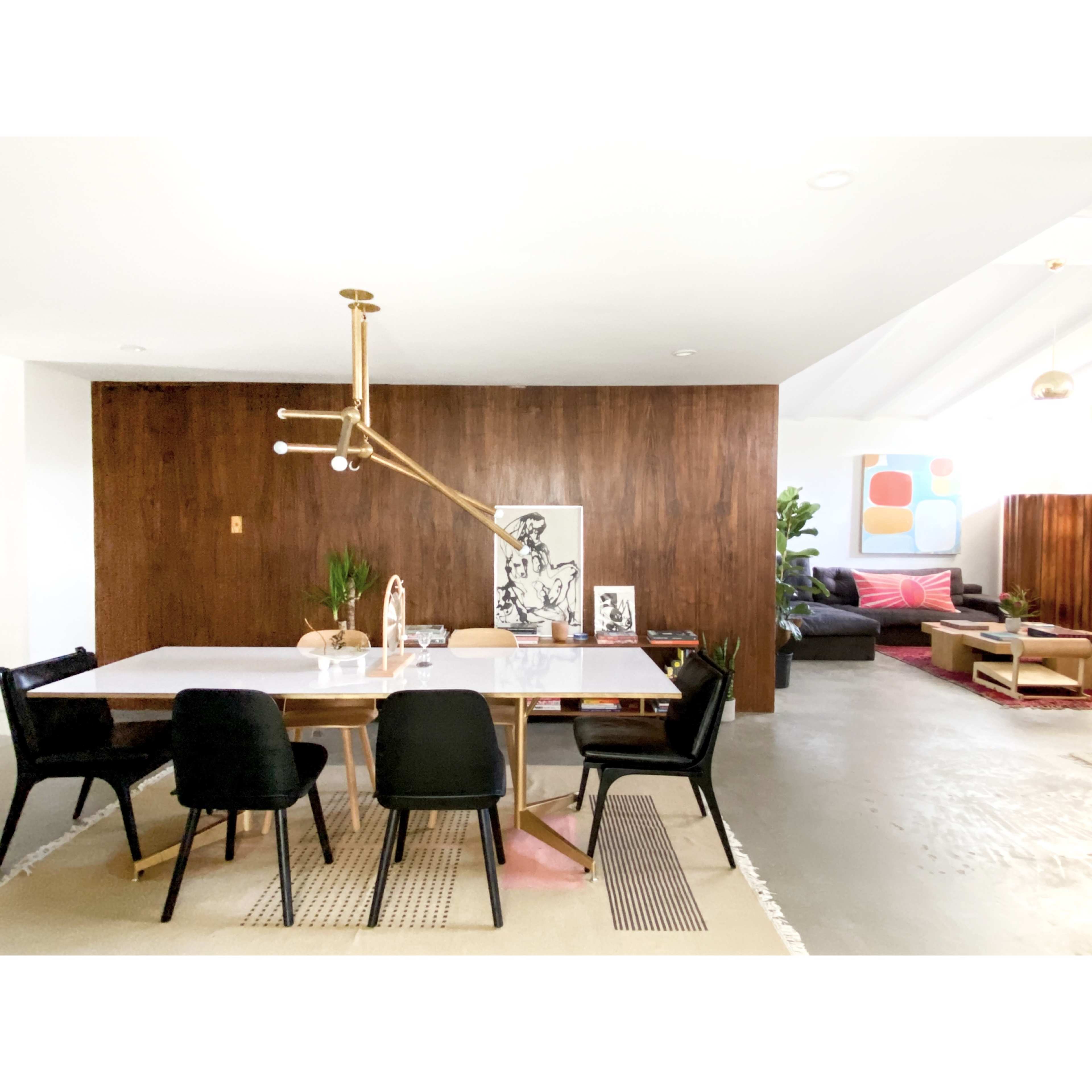A modern dining area features a marble table surrounded by black chairs, with a wood-paneled wall in the background and an adjacent living space visible.