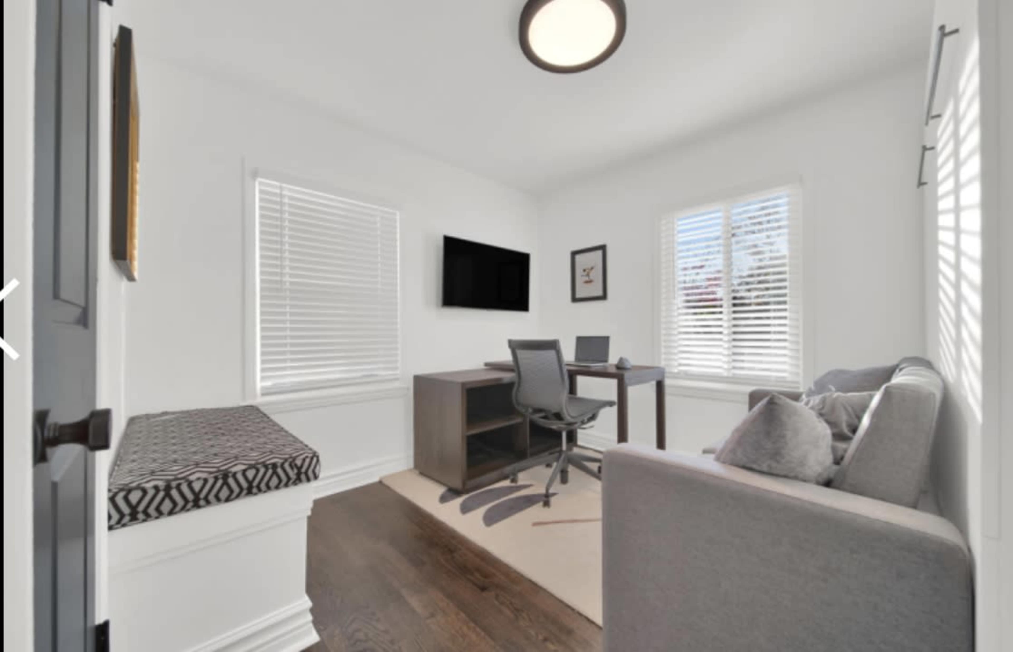 A bright home office features a desk with two chairs, a television mounted on the wall, and a seating area with a sofa by the window.