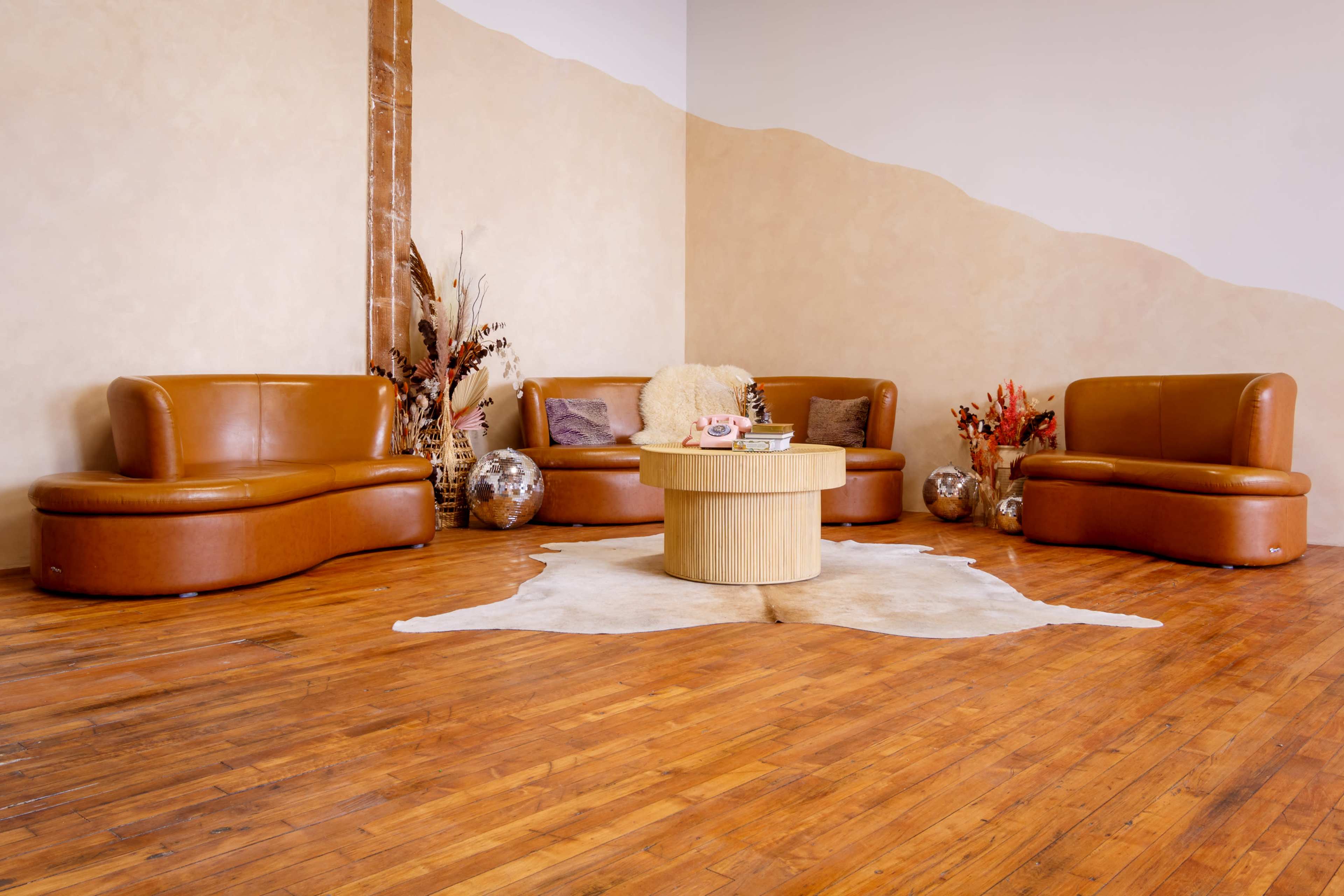 A circular wooden coffee table surrounded by curved brown leather couches on a wooden floor, with decorative elements like vases and plants in the background.