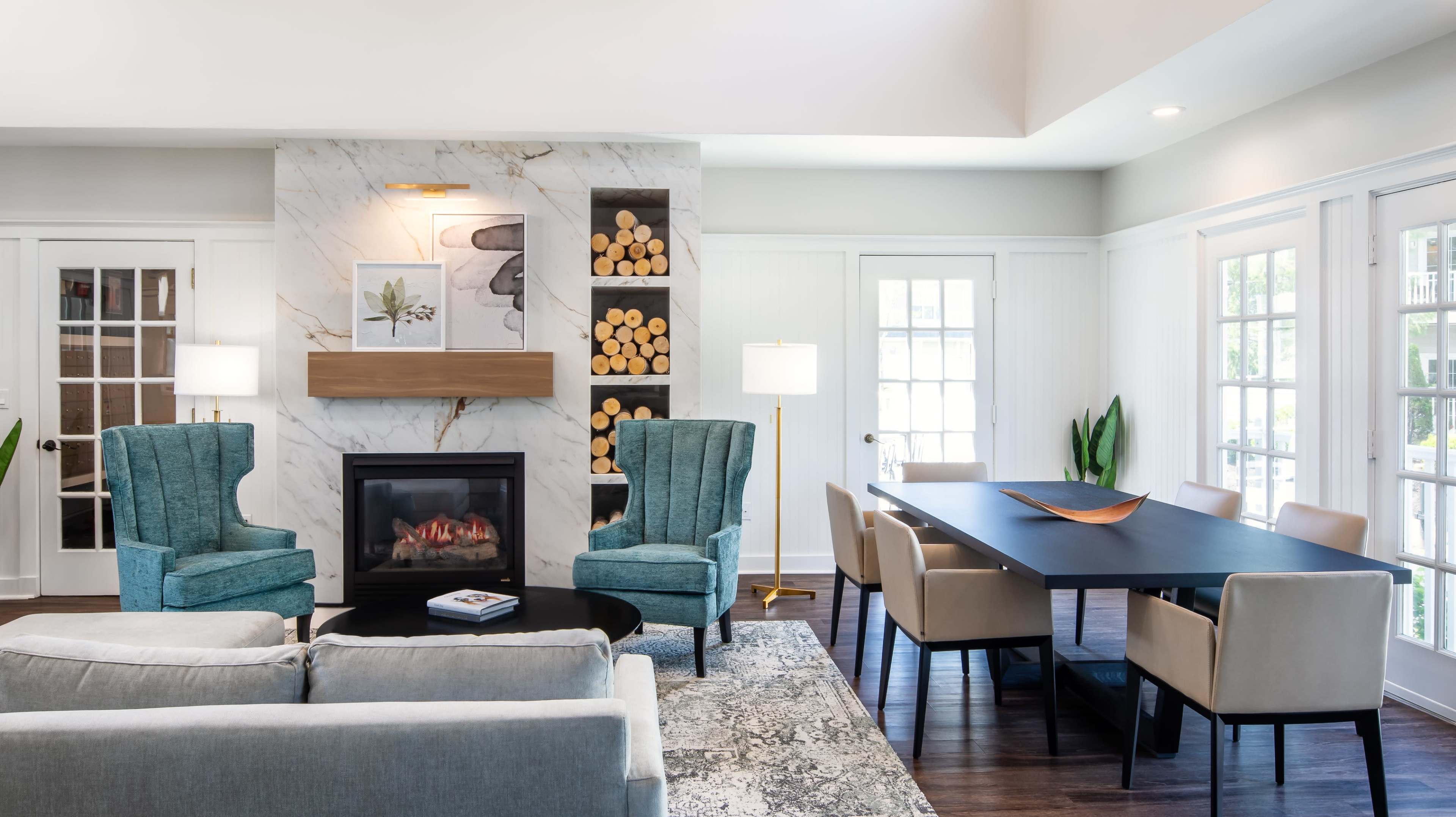 A modern living room featuring a fireplace with decorative logs, two blue armchairs, a gray sofa, a dining table with beige chairs, and large windows allowing natural light.