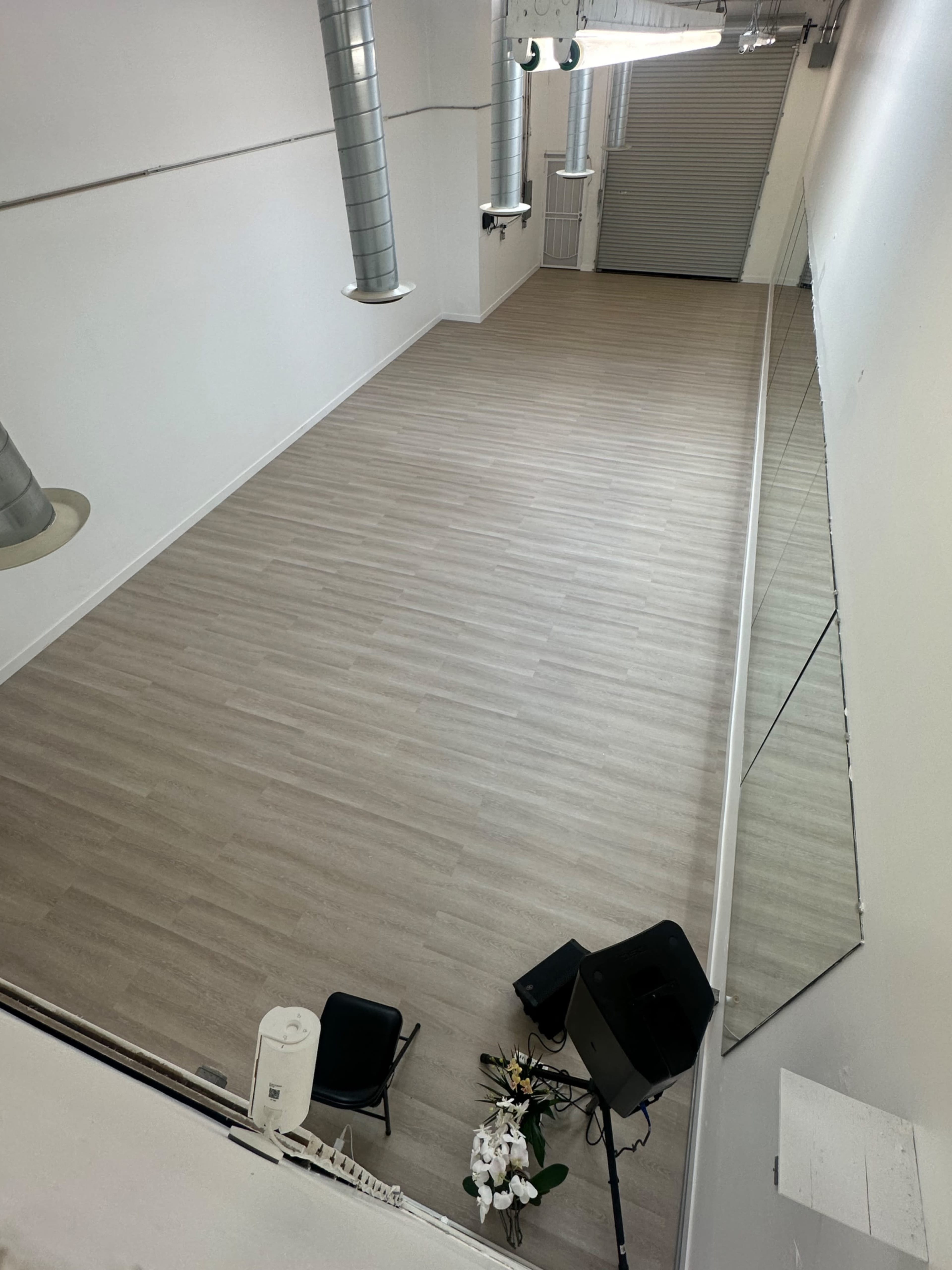 The image shows a large, empty studio space with light-colored flooring, a wall of mirrors, and a speaker setup in the corner.