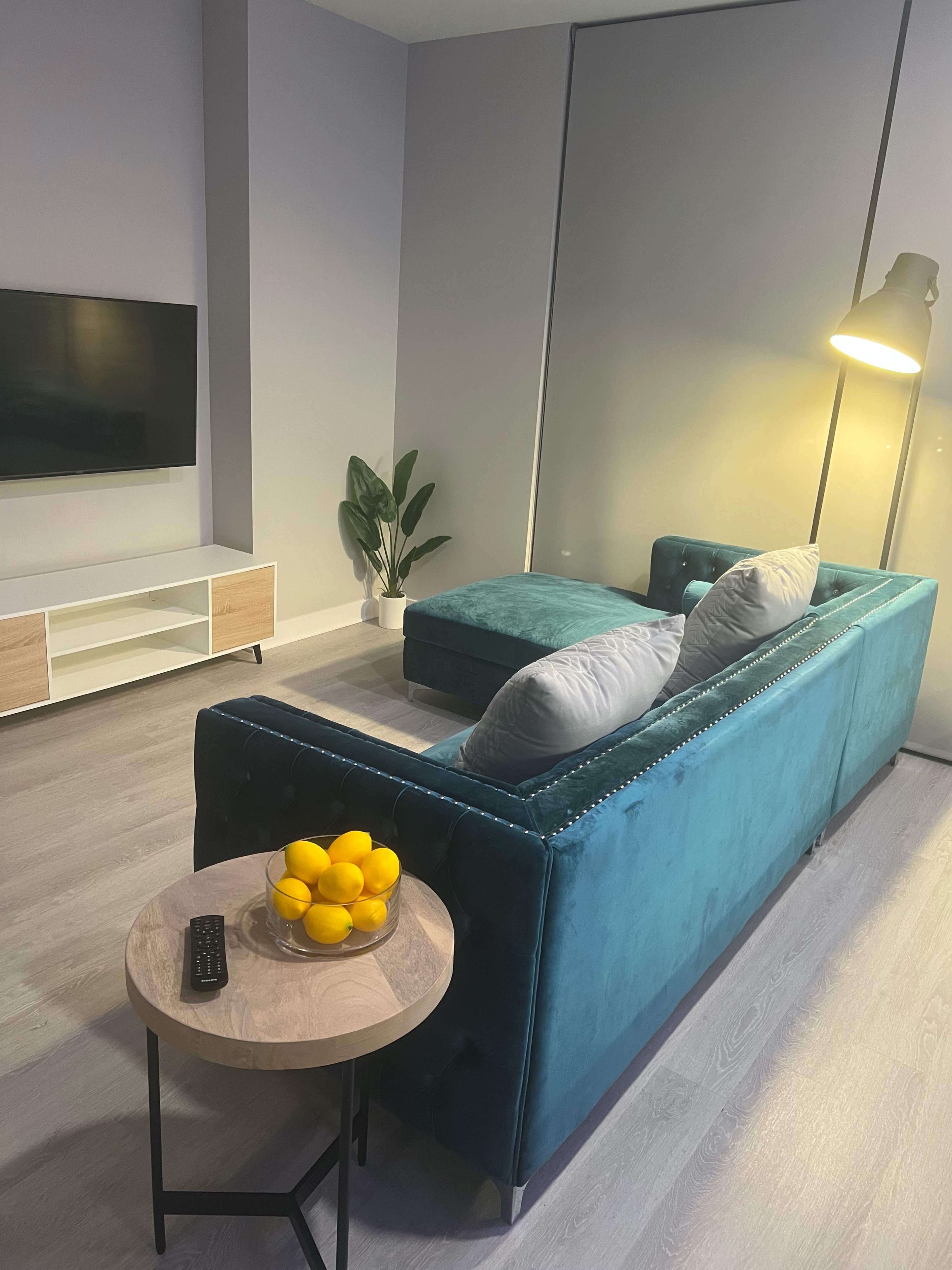 A teal sofa with gray pillows is positioned next to a floor lamp and a small round table holding a bowl of lemons in a modern living room.