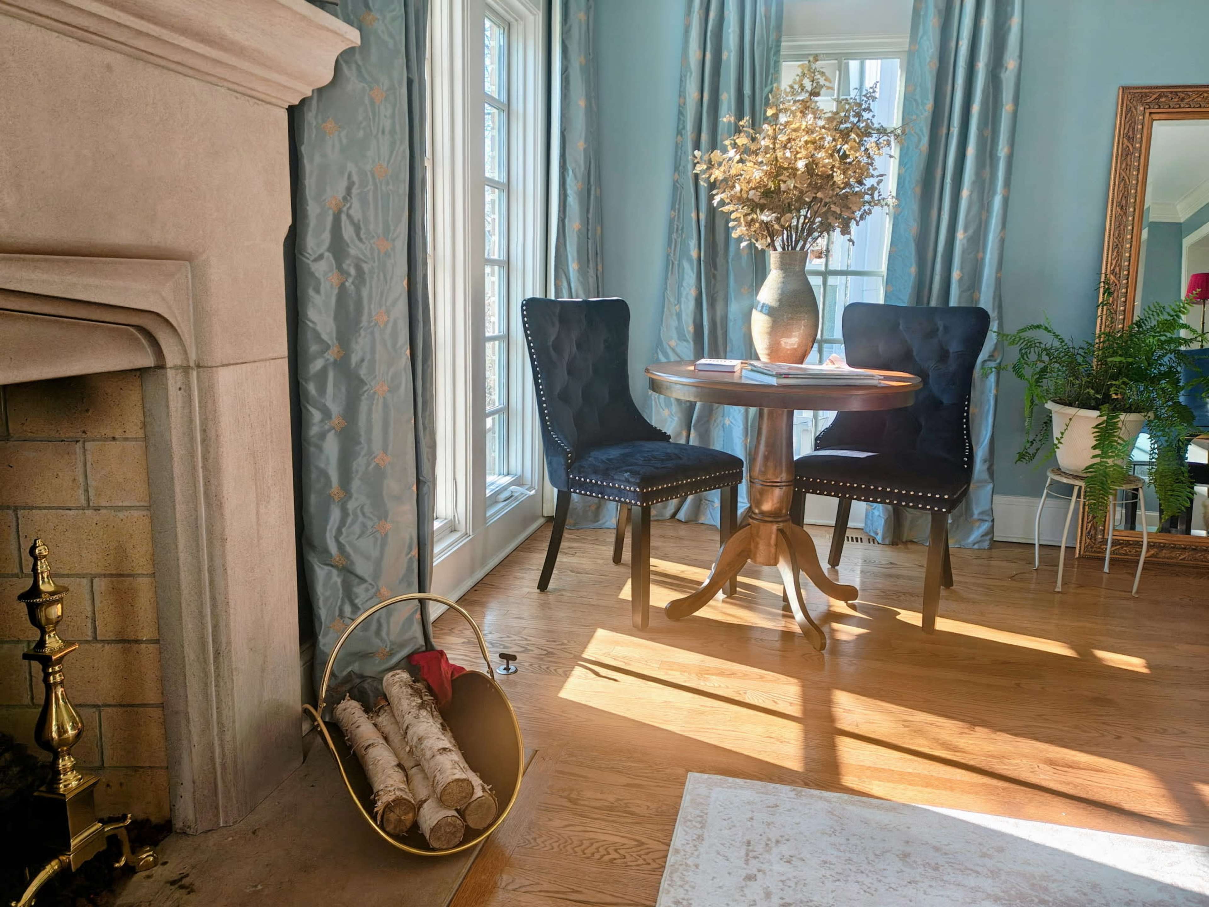 A small round table with two dark chairs is set beside a fireplace, adorned with a vase of dried flowers and illuminated by sunlight filtering through the window.
