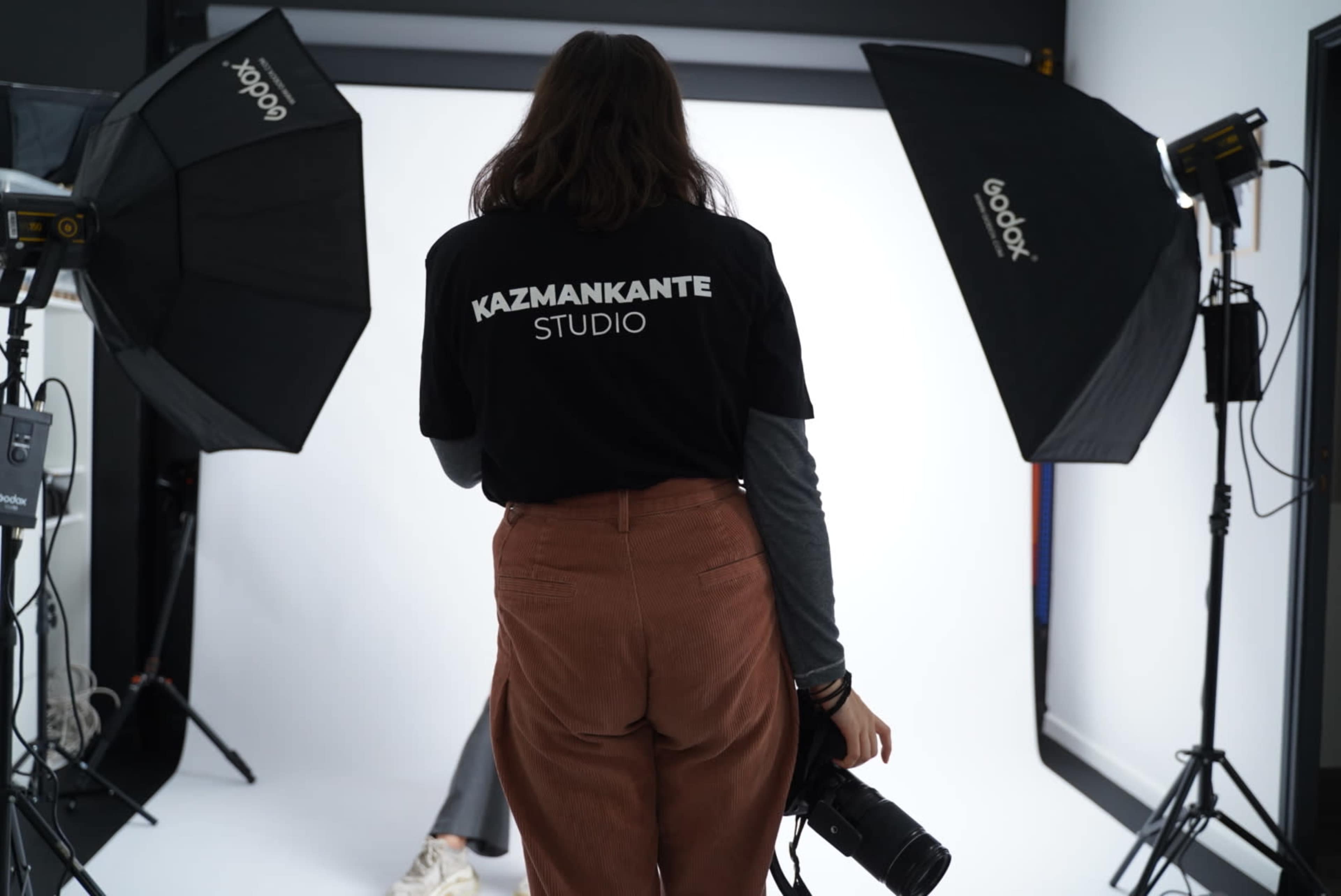 A person stands in a photography studio wearing a shirt that says "KAZMANKANTE STUDIO" while holding a camera, with lighting equipment set up around them.
