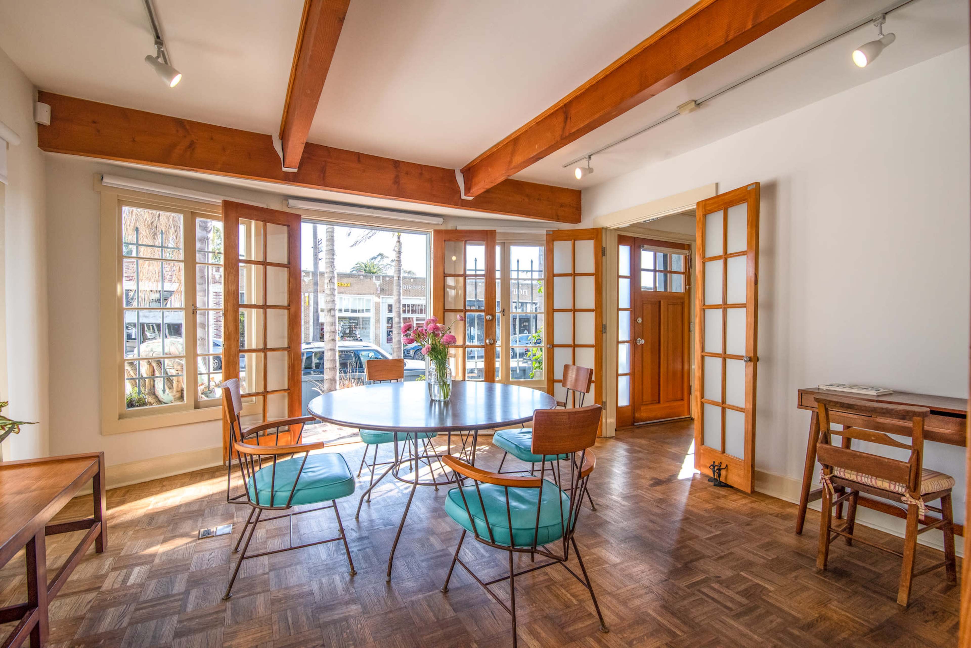 A dining area features a round glass table surrounded by wooden chairs, with multiple large windows letting in natural light.