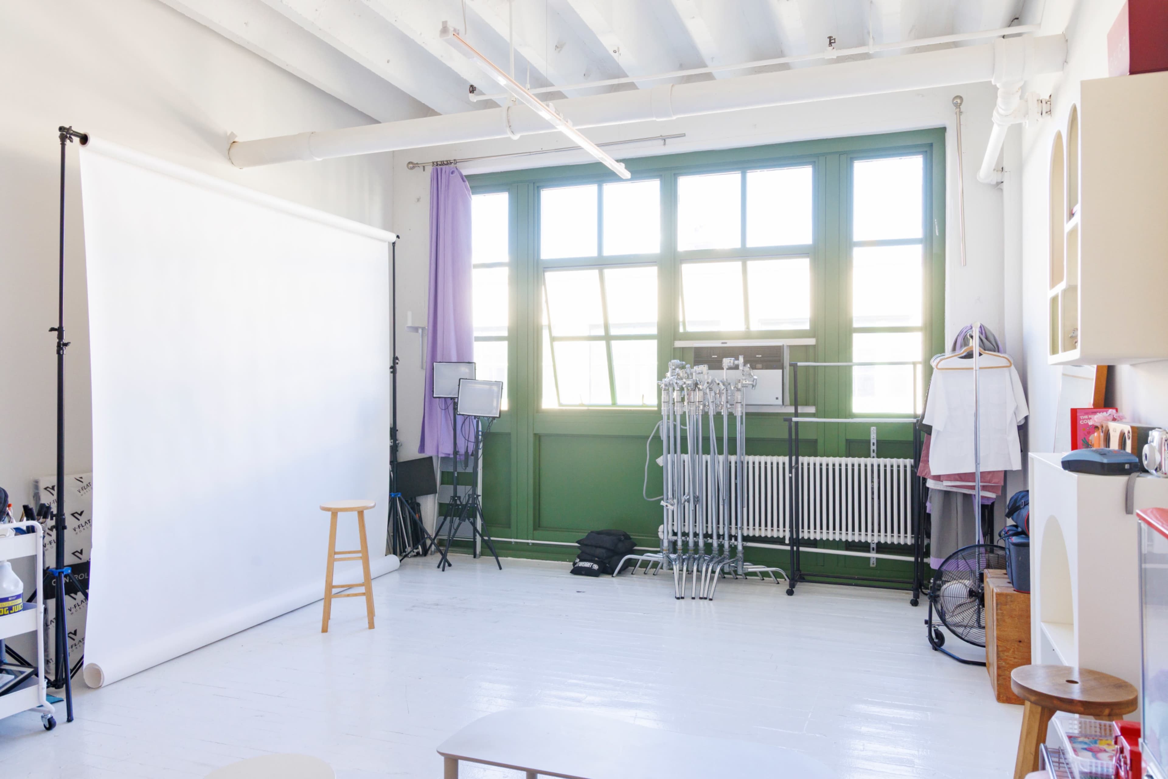 The image shows a bright, empty photography studio with a white backdrop, a wooden stool, and a large window letting in natural light.
