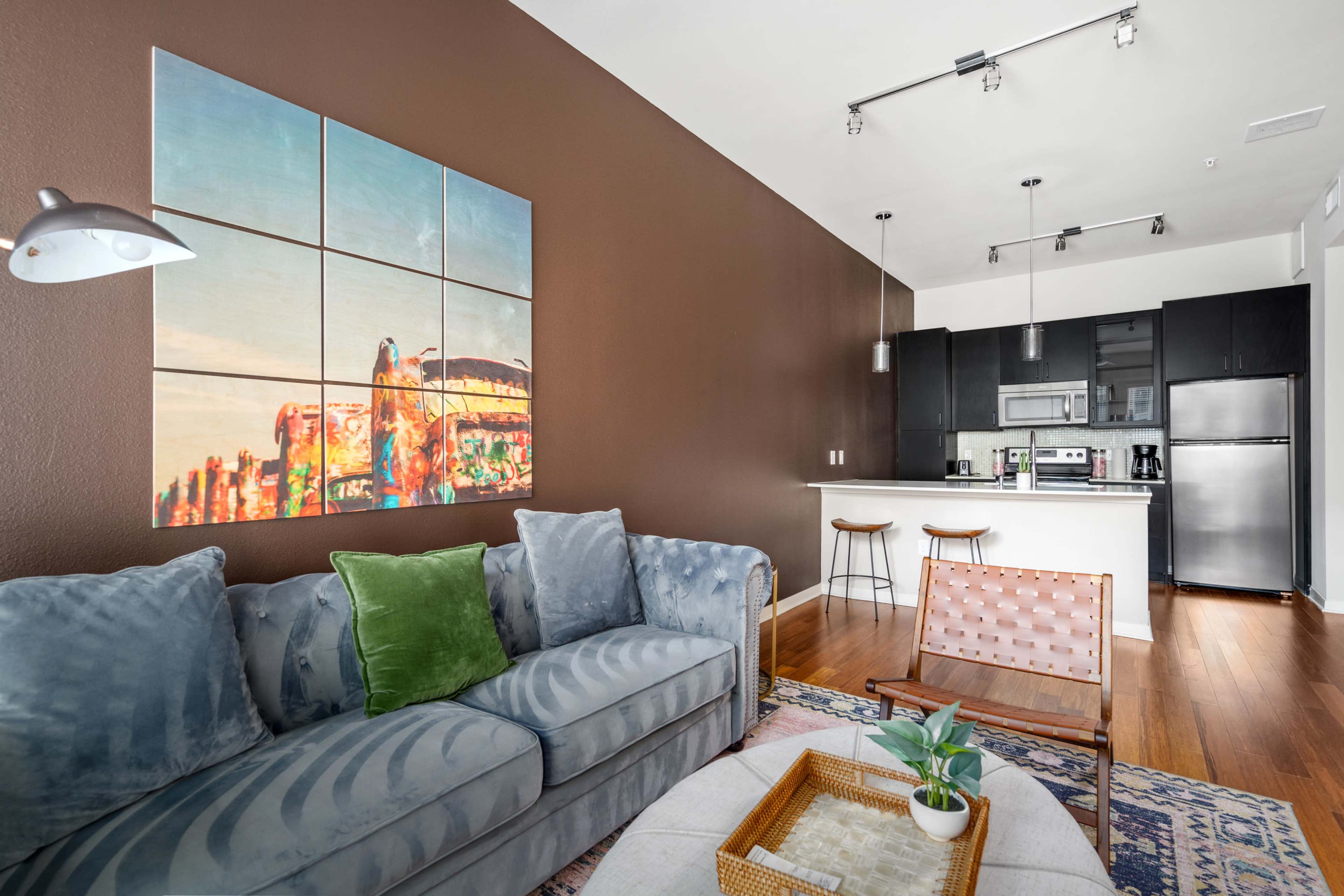 A modern living room with a blue sofa, a brown accent wall featuring a multi-panel artwork, and a kitchen area in the background.