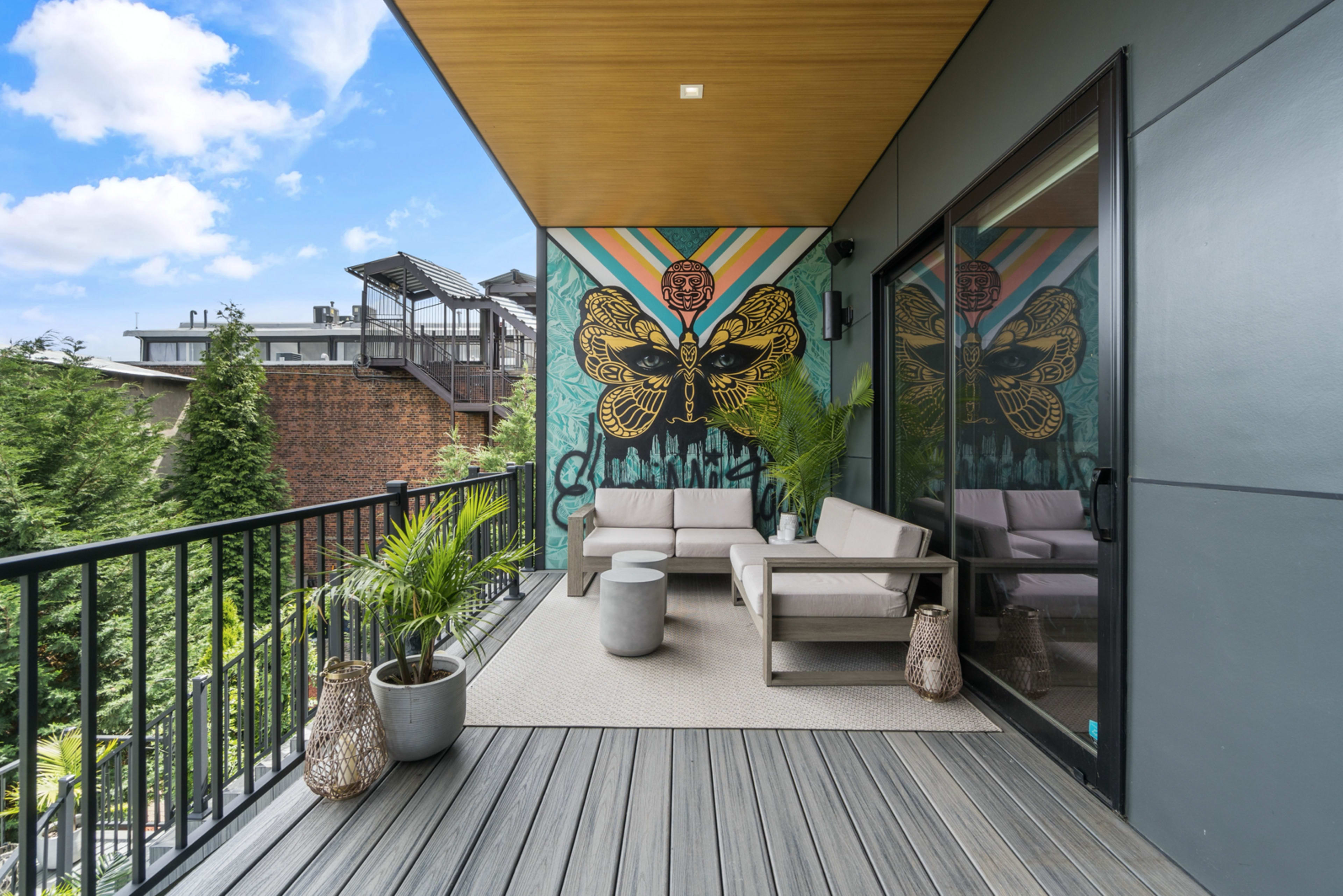 A balcony with modern furniture, featuring a mural of a butterfly on the wall and surrounded by potted plants.