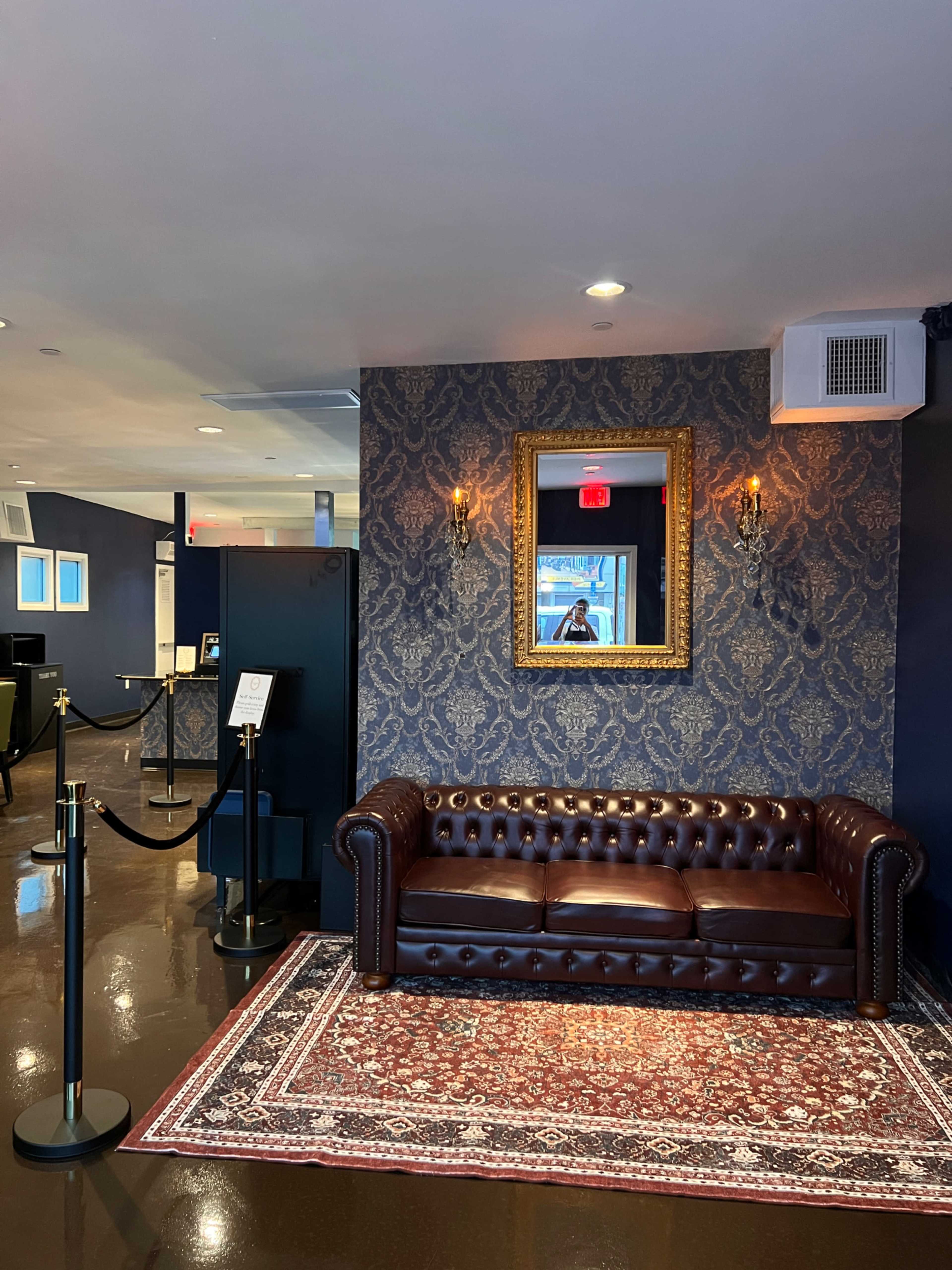 The image shows a lobby area featuring a brown leather chesterfield sofa, ornate wallpaper, a decorative mirror, and a patterned rug, with a stanchion barrier visible in the foreground.