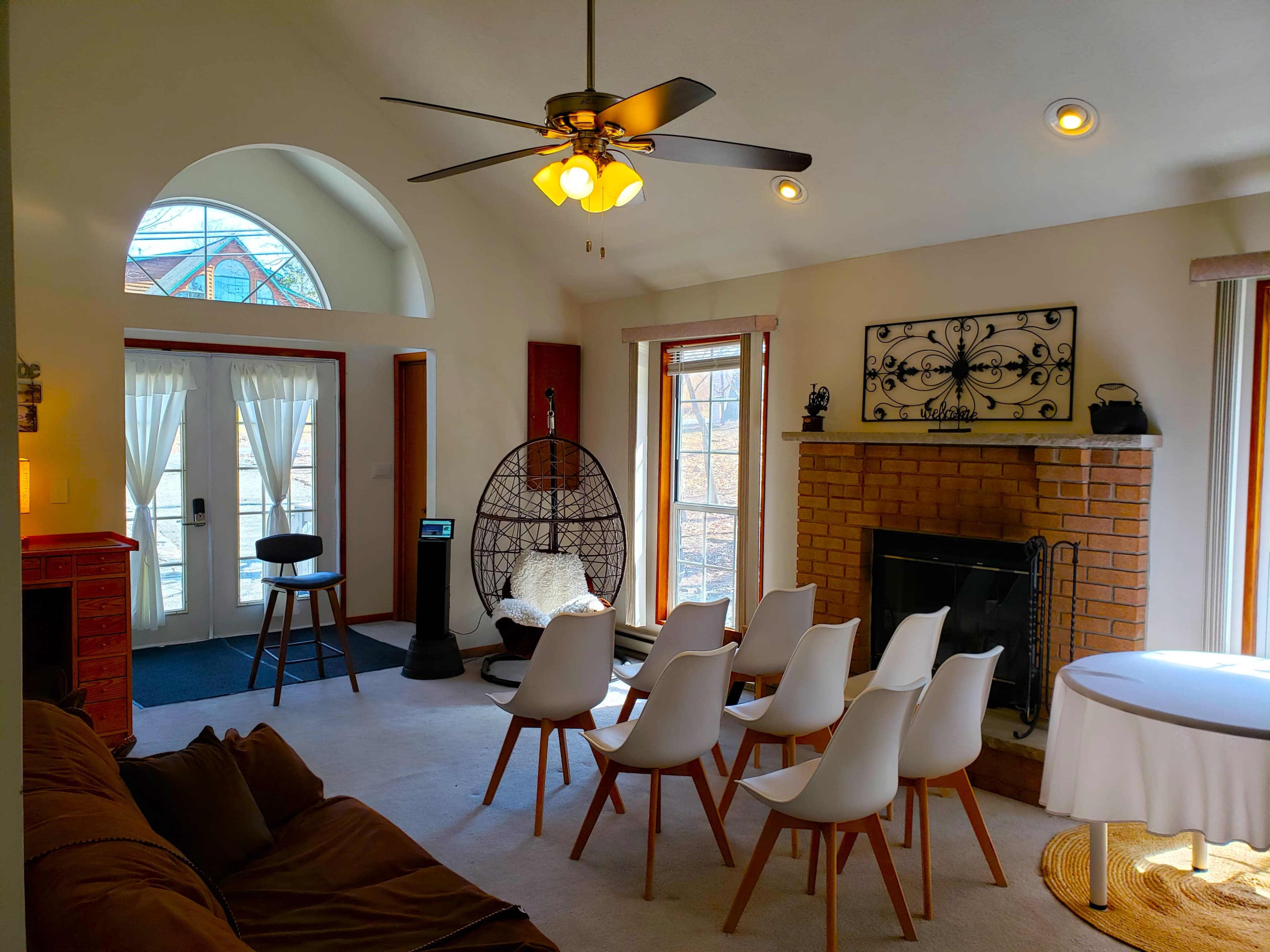 The image shows a living room with a circular seating arrangement of white chairs, a fireplace, and sunlight streaming through large windows.
