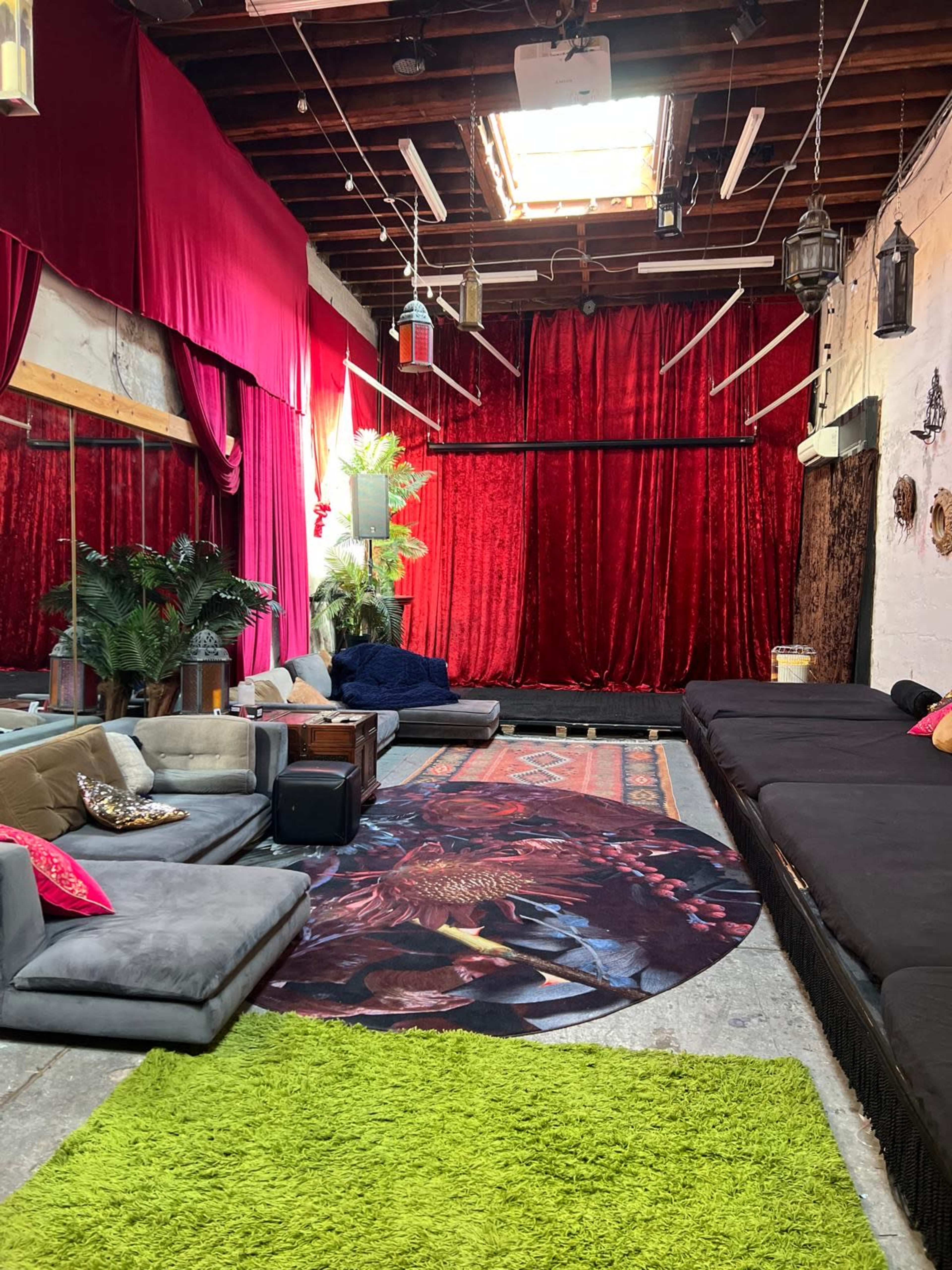 The image shows a spacious indoor area with plush seating, a large green rug, and red velvet curtains draping the walls, illuminated by natural light from a skylight.
