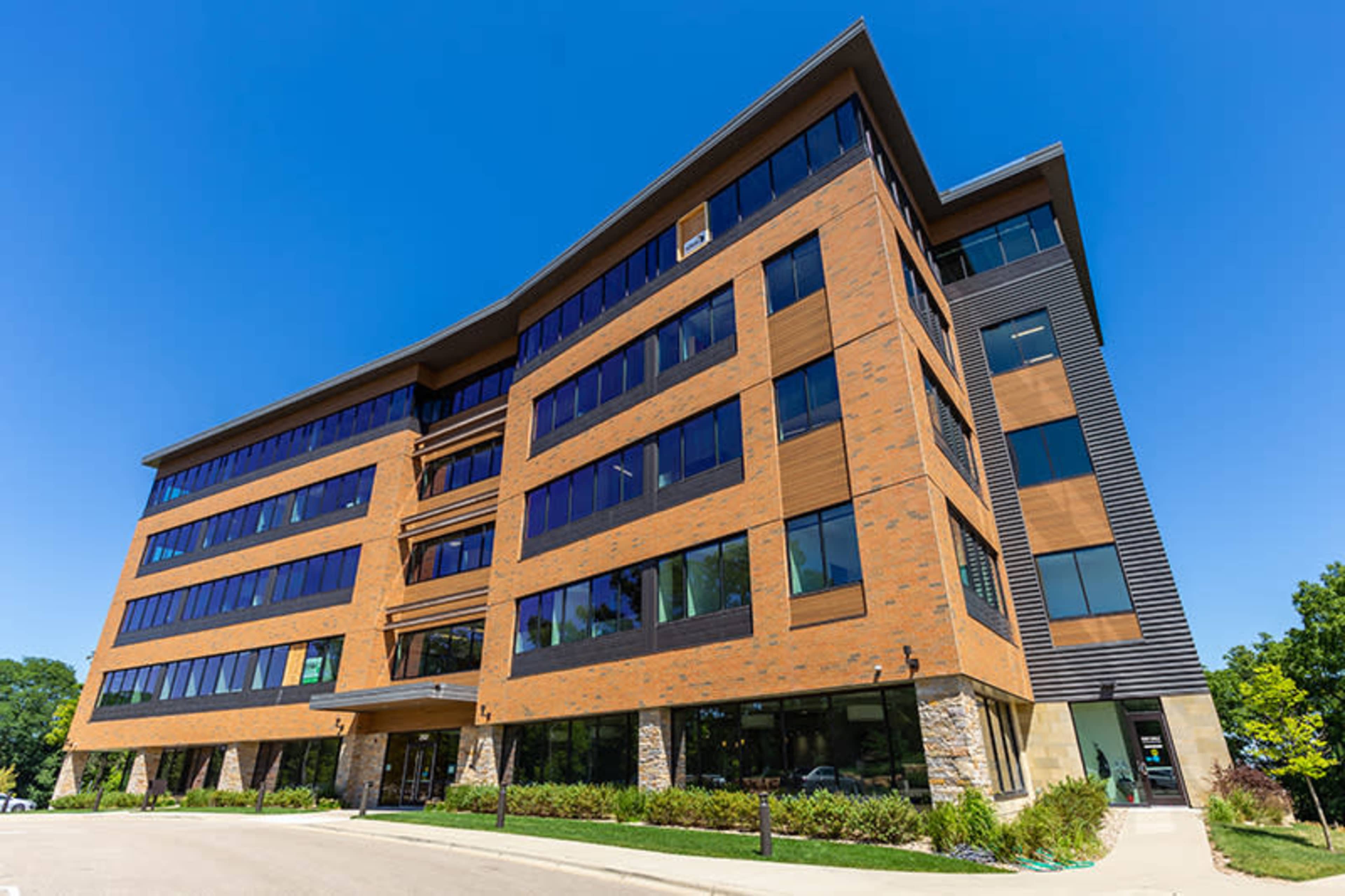 A modern, five-story office building features a mix of brick and wood paneling, large windows, and landscaped surroundings.