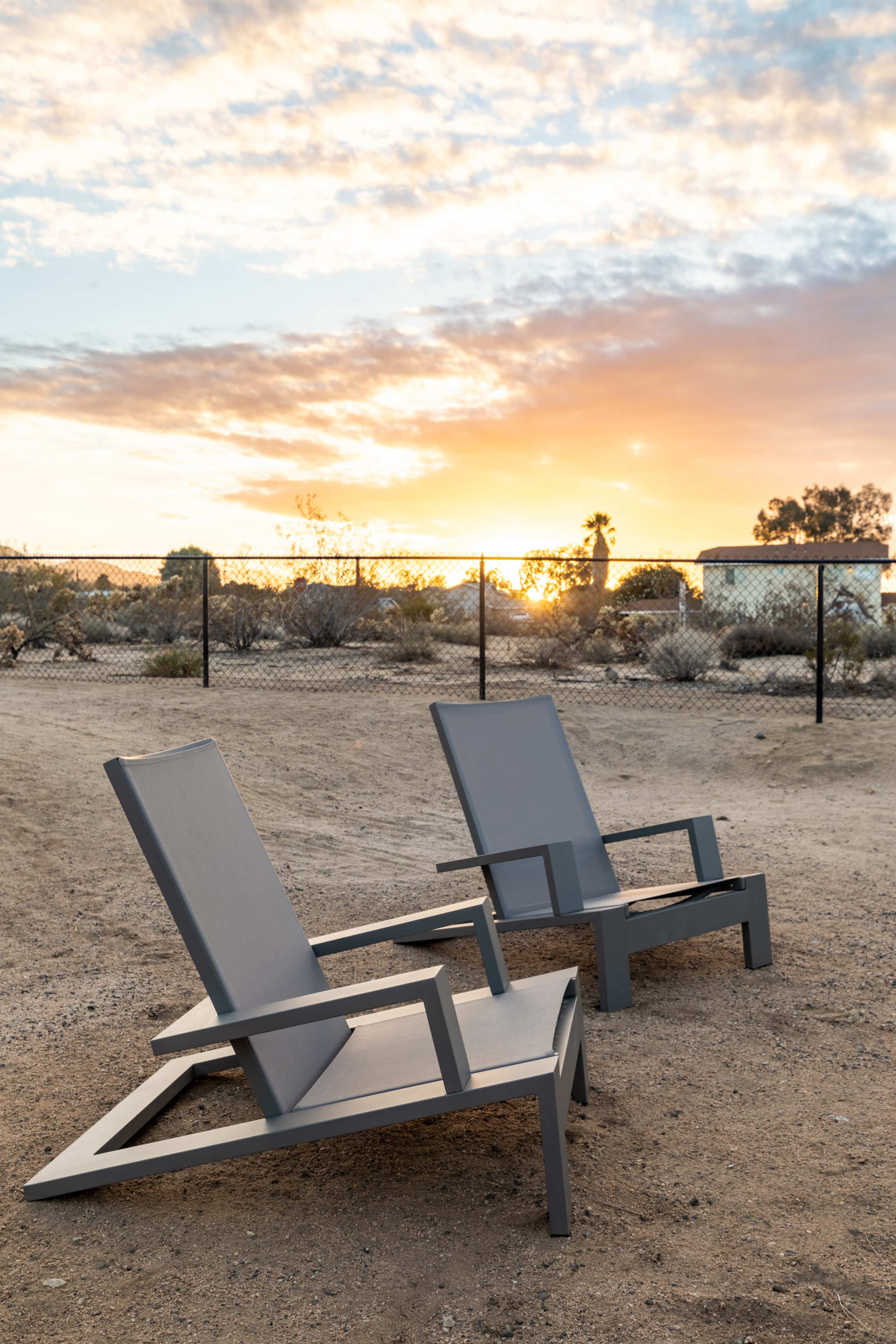Two modern lounge chairs are positioned on a sandy area facing a sunset, with a fence and desert vegetation in the background.