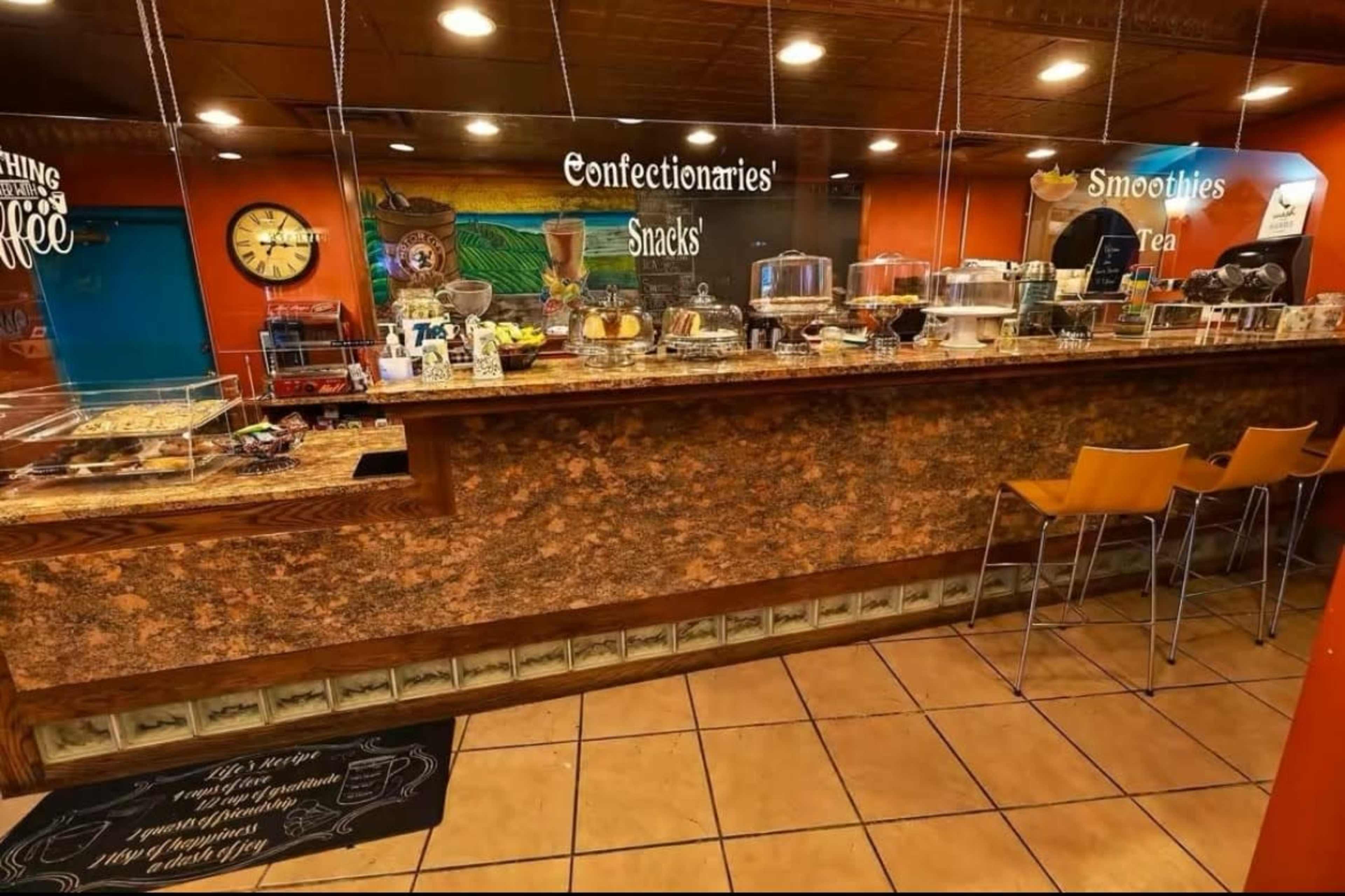 The image shows a bakery café counter with a variety of snacks and beverages displayed behind a glass countertop, along with seating in front.