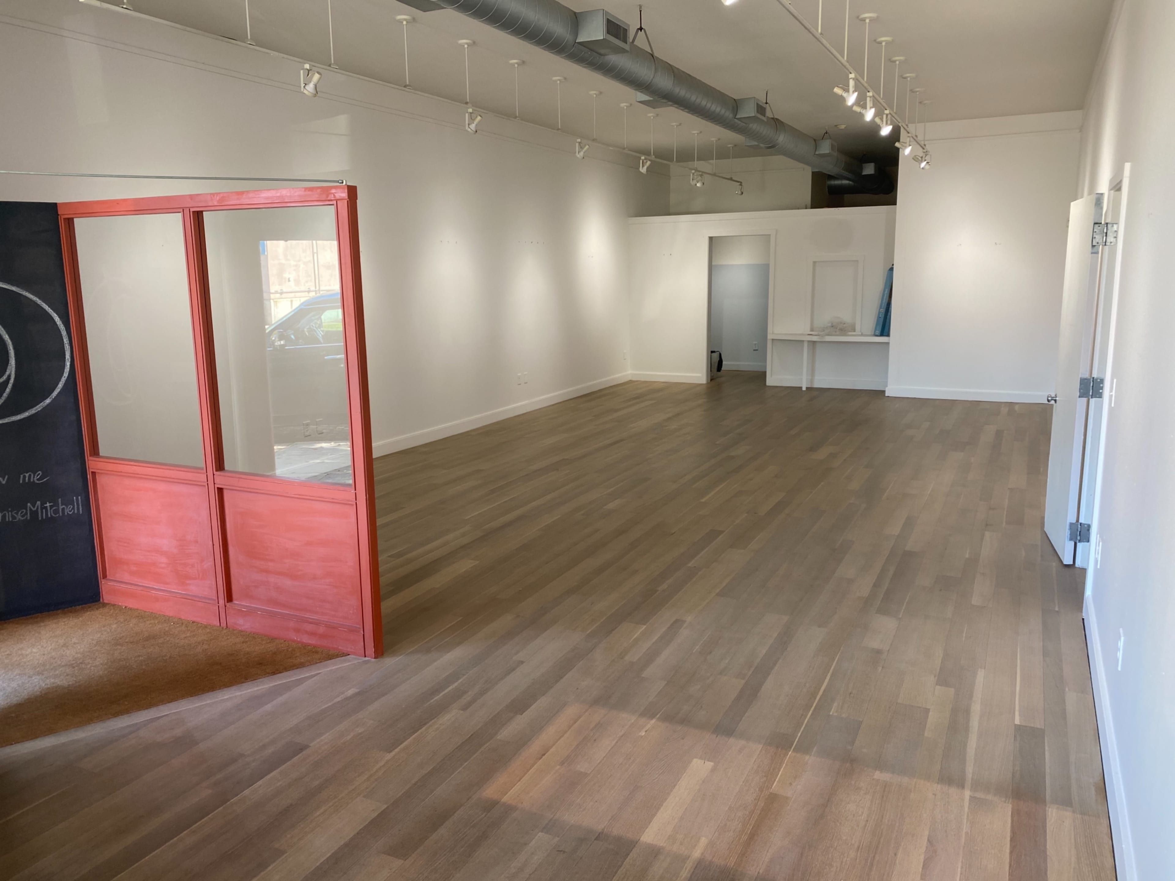 An empty commercial space with wooden floors, white walls, and a glass-paneled door leading to another room.